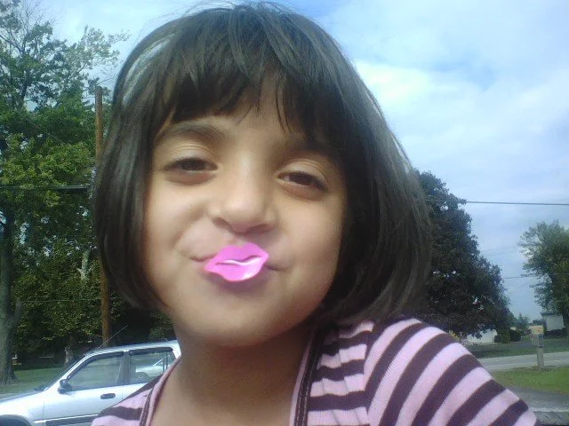 Young girl with short dark hair making a kissing face with pink lip-shaped objects on her lips, outdoors with trees and a car in the background.