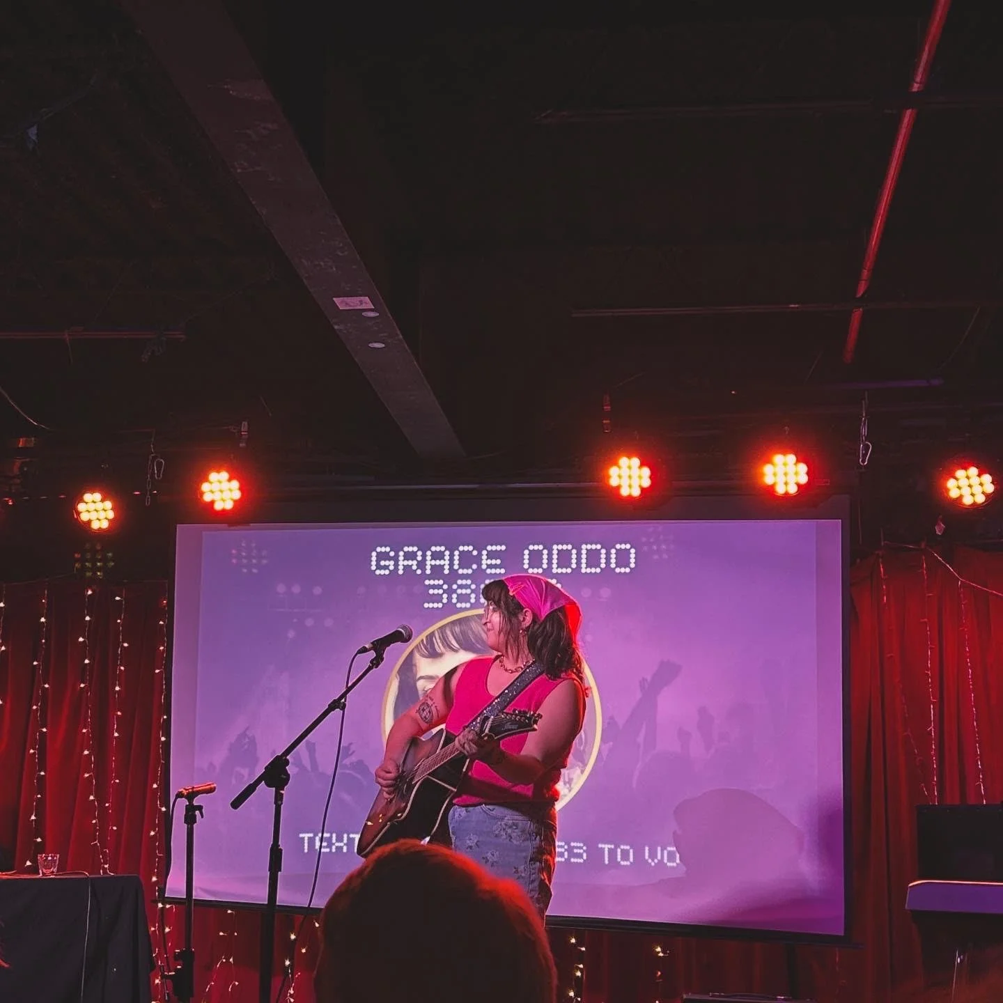 Performer Grace Odoo on stage playing guitar with a pink headscarf, microphone, colorful background with text and red curtains.
