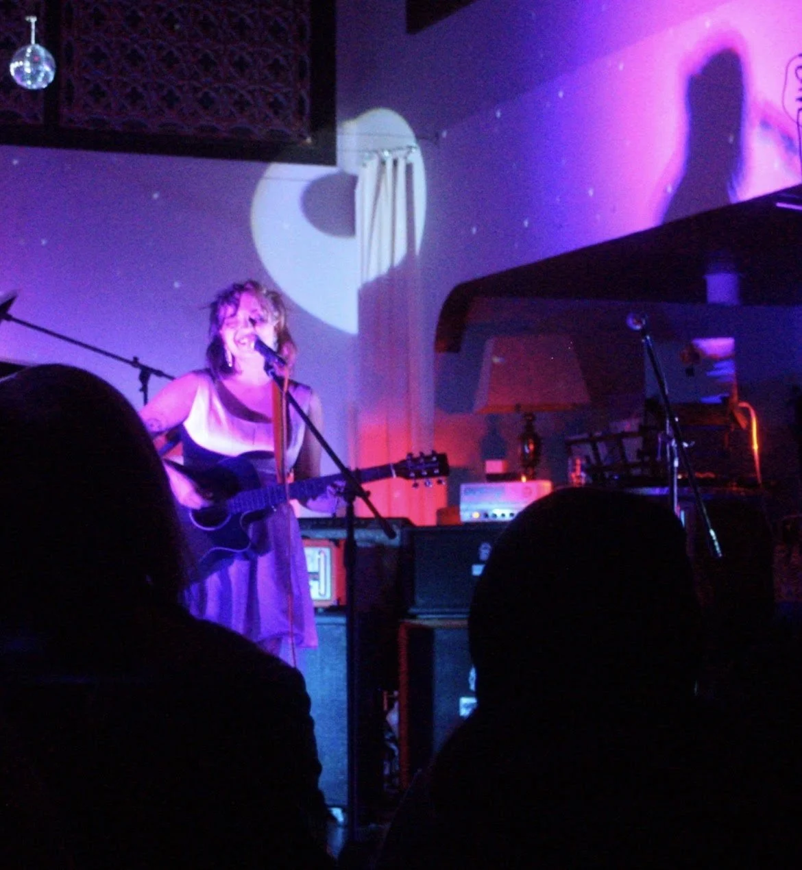 A woman singing and playing guitar on stage in a dimly lit venue with purple and pink lighting, audience silhouettes in the foreground, and a disco ball hanging from the ceiling.