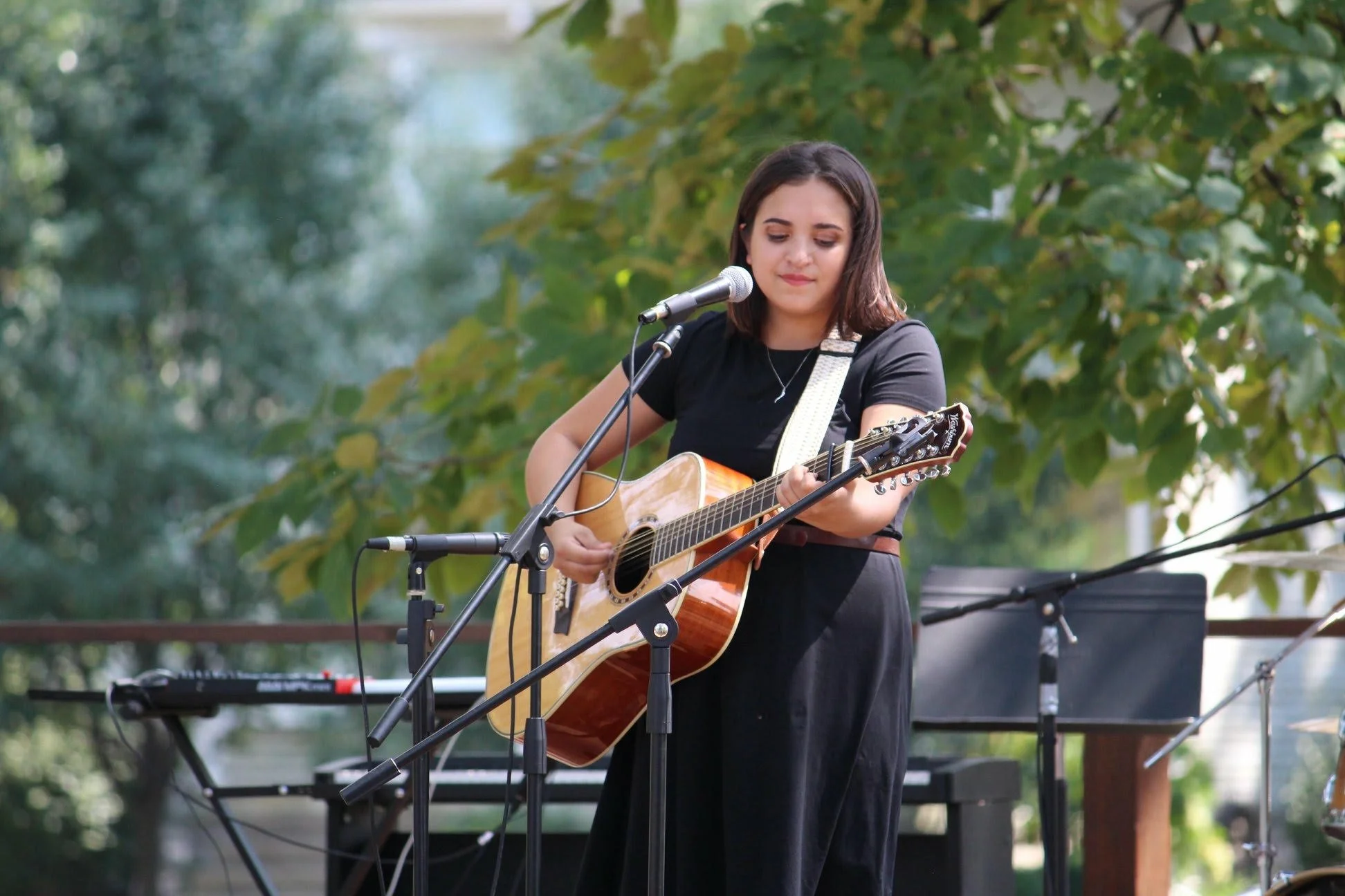 A young woman with shoulder-length brown hair playing an acoustic guitar and singing into a microphone outdoors during daytime, with green trees in the background.