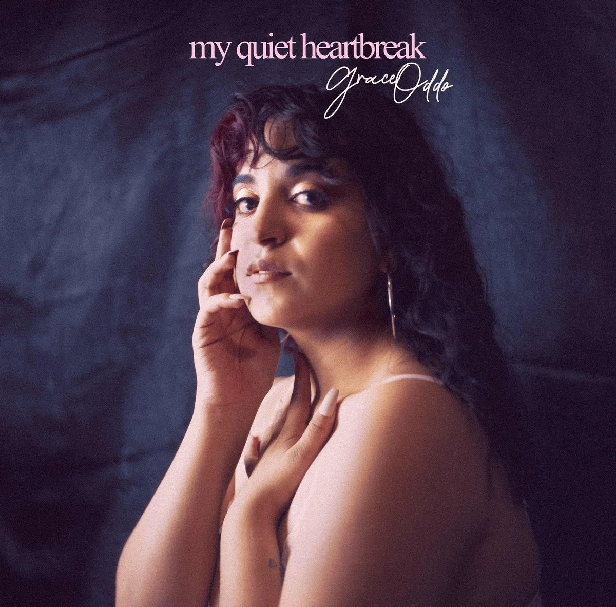 Cover art for Grace Oddo's album titled "My Quiet Heartbreak," featuring a woman with dark, wavy hair, looking at the camera with her hand near her face, against a dark background.