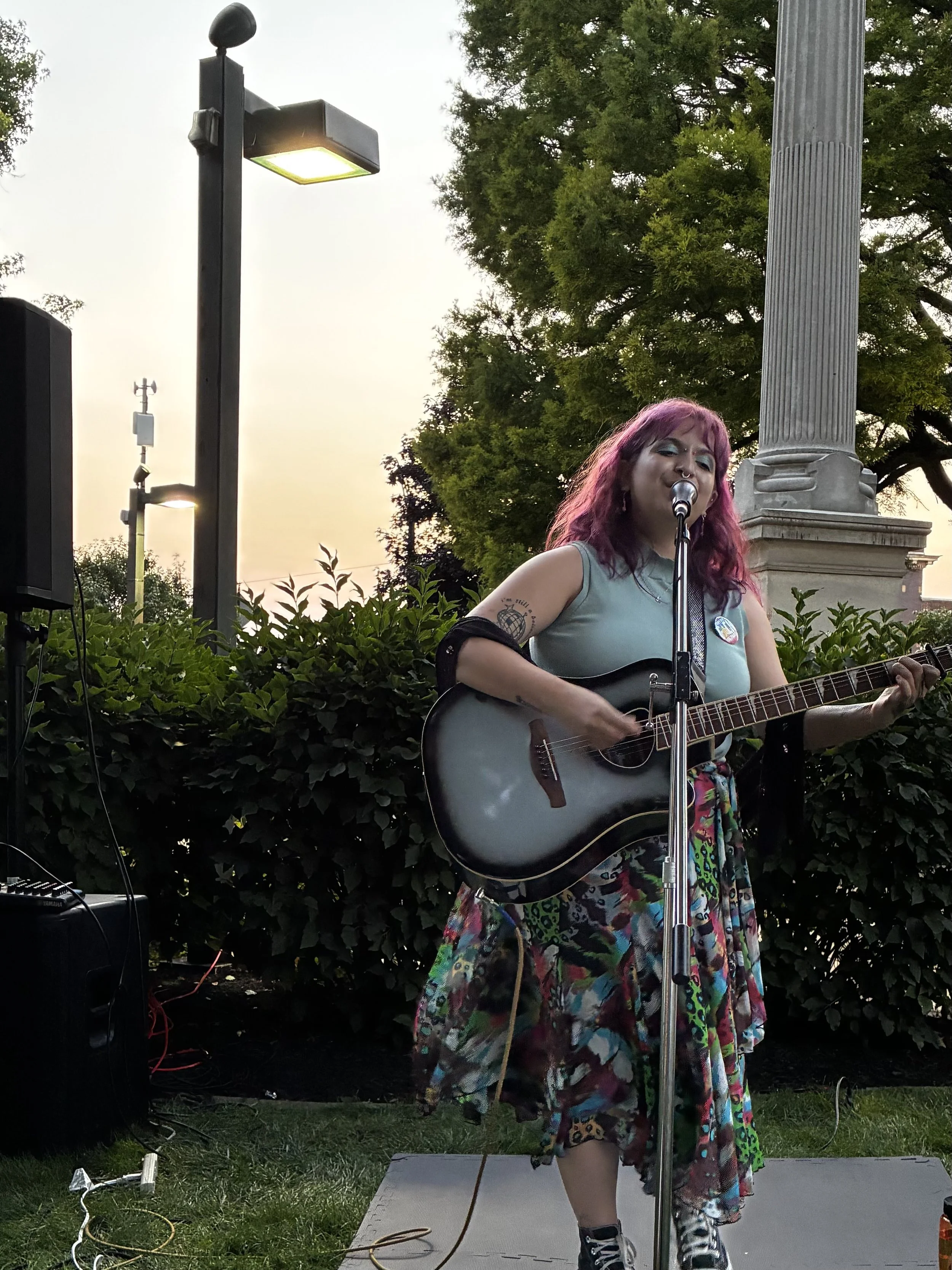 A woman with pink hair singing and playing guitar outdoors at sunset. She is standing on a small stage with a microphone, in front of green bushes, with a large column and trees in the background.