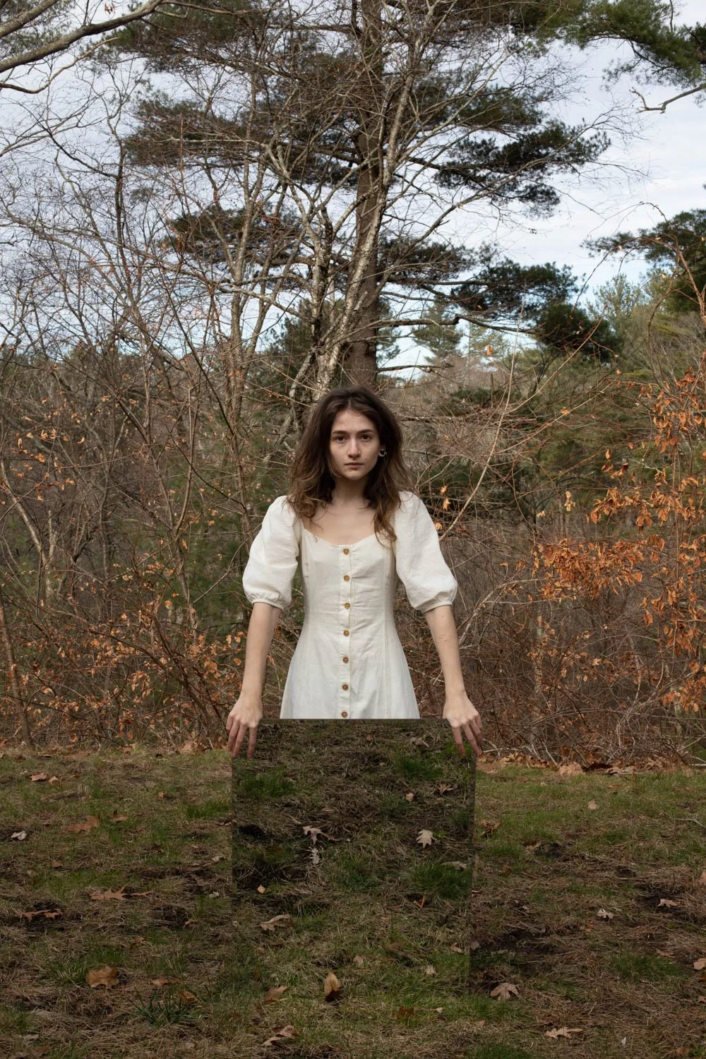 A young woman with brown hair wearing a white dress with buttons stands outdoors among leafless and orange foliage, with a large tree in the background.