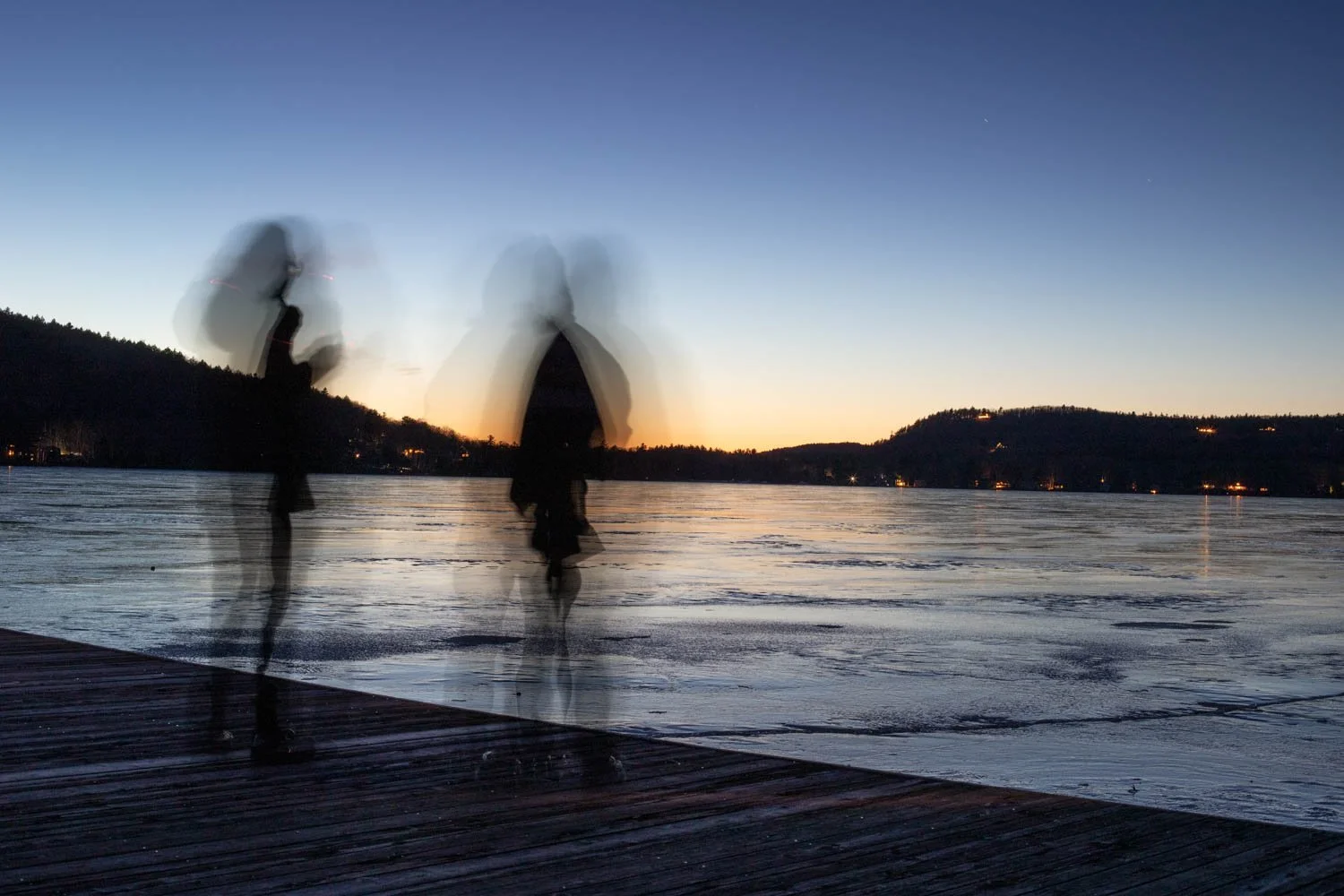 People walking on a wooden dock by a frozen lake at sunset, with hills in the background and motion blur indicating movement.