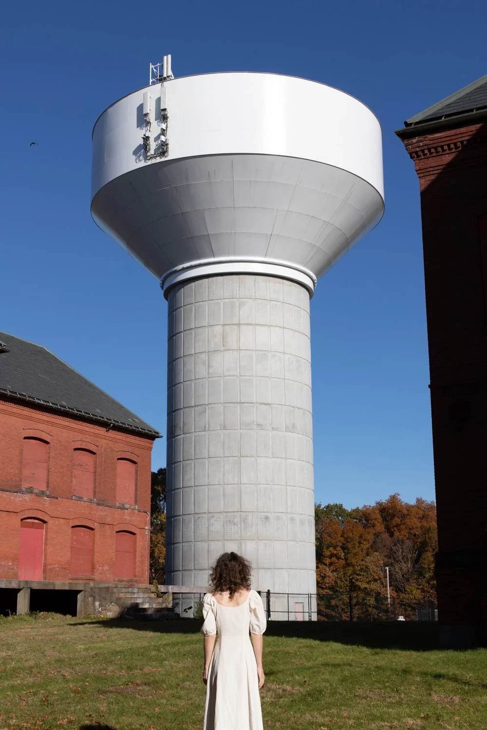 A woman in a white dress standing outside, looking at a large white water tower between two brick buildings under a clear blue sky.