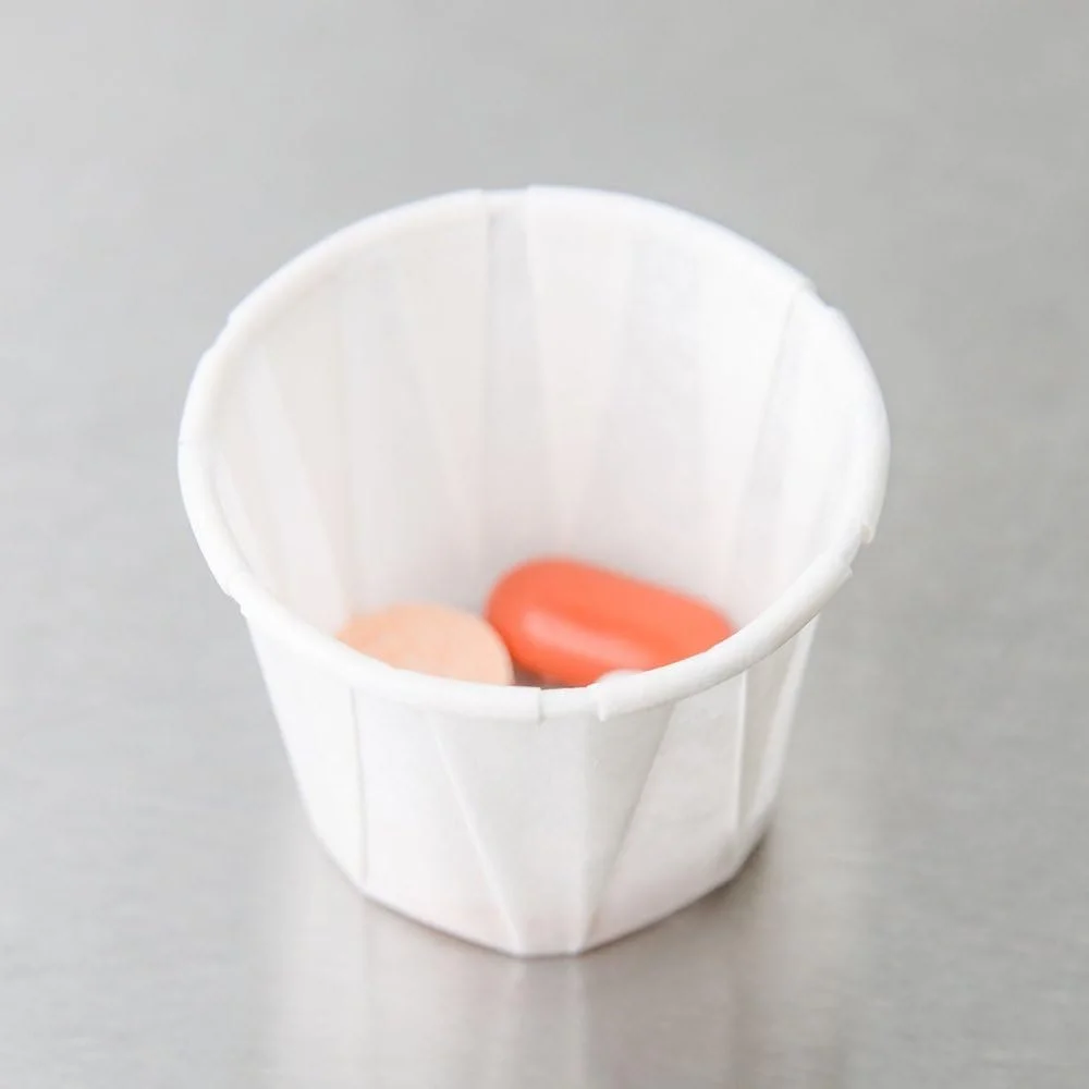 A white paper cupcake liner with two pink and orange pills inside, placed on a silver surface.