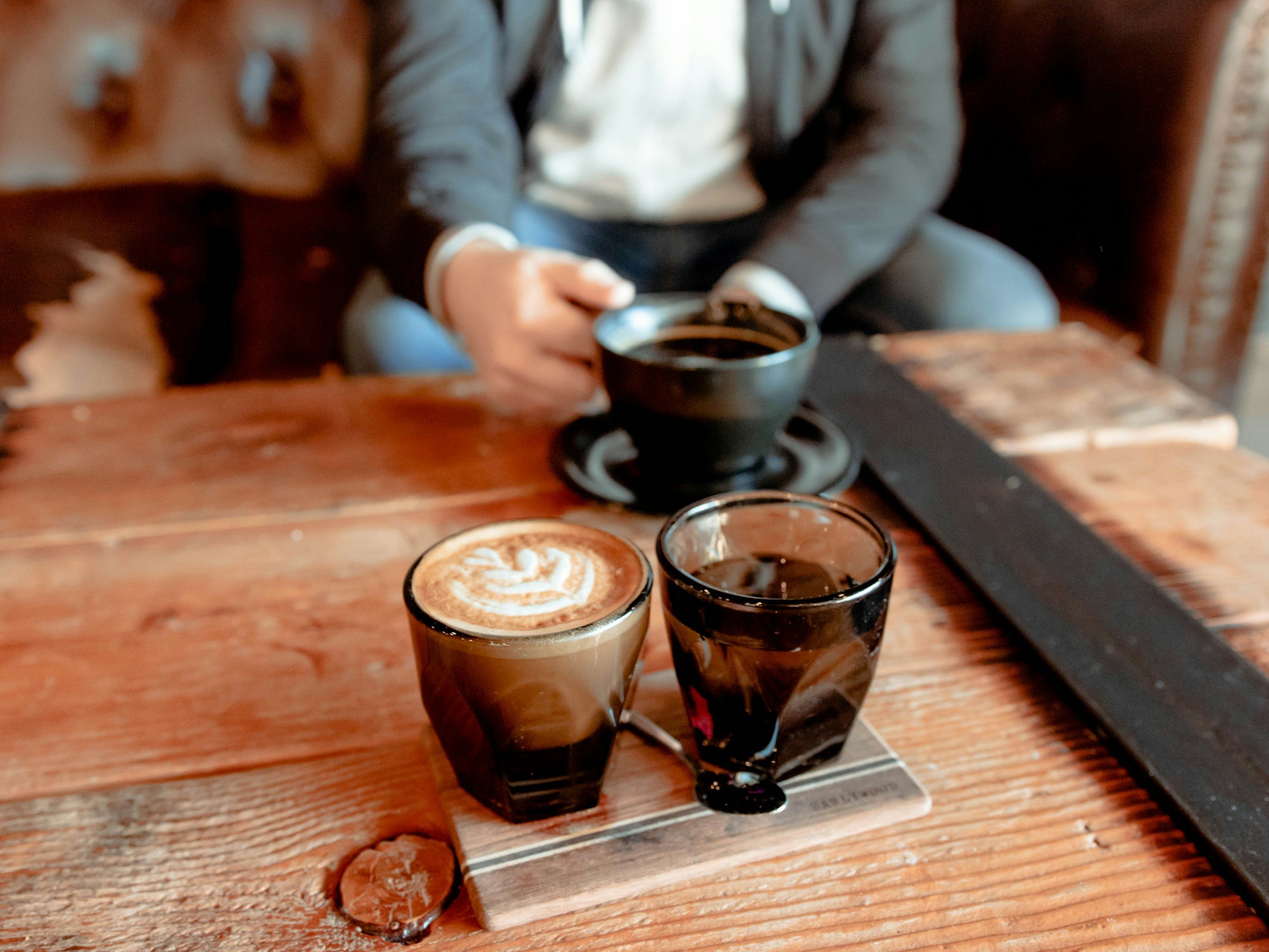 Three glasses of coffee with different coffee art placed on a wooden table, with a person in a dark suit and white shirt sitting at the table in the background.