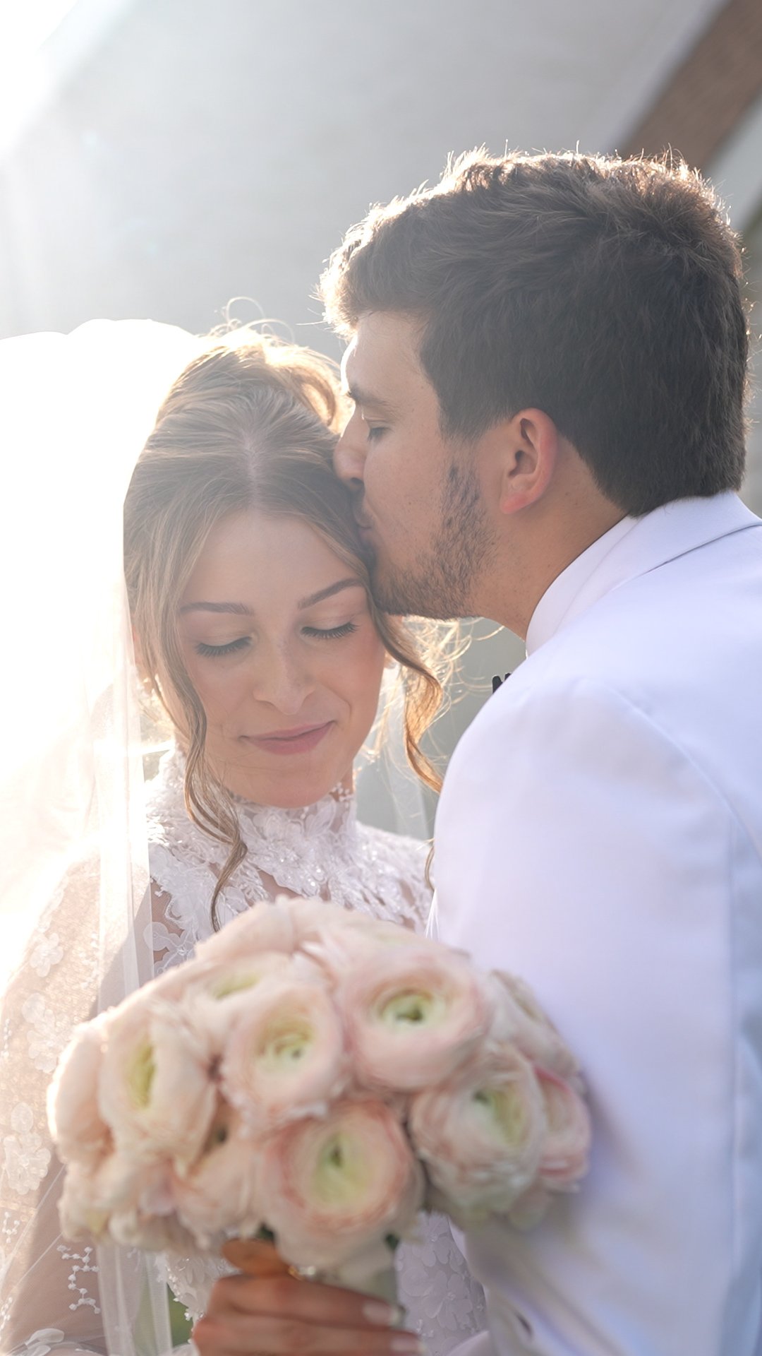 A bride and groom sharing a kiss during their wedding, with the bride holding a bouquet of pink and white flowers.