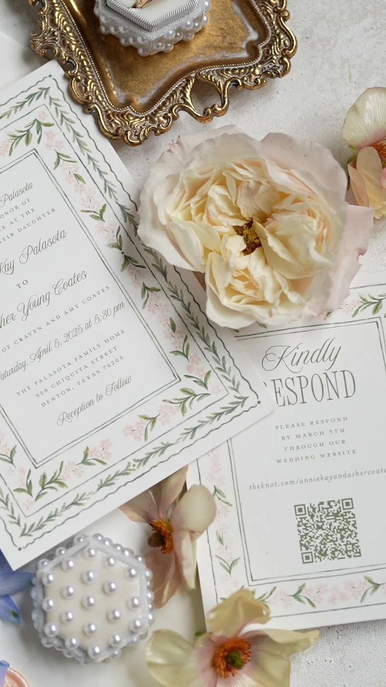 Wedding invitation surrounded by cream-colored flowers and decorative items on a white surface.