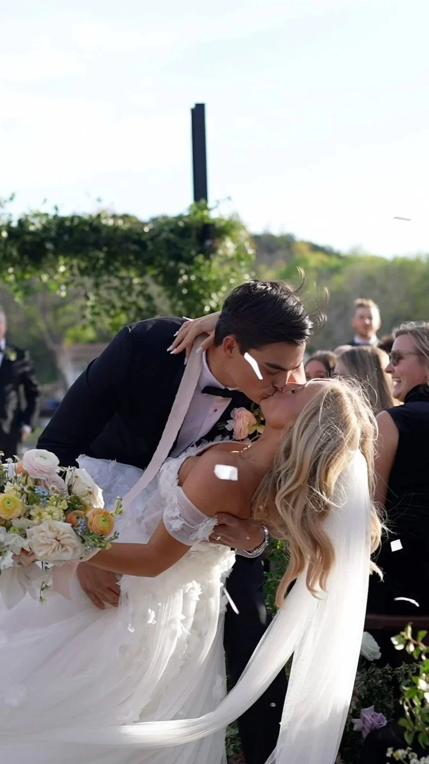 A newlywed couple sharing a kiss during their wedding ceremony, surrounded by friends and family, with sunlight and greenery in the background.