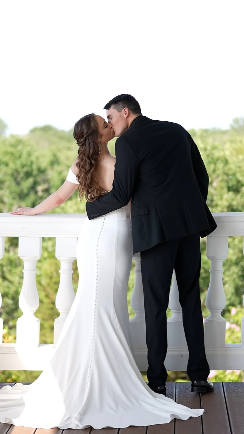 A couple in wedding attire kissing on a balcony with lush greenery in the background.