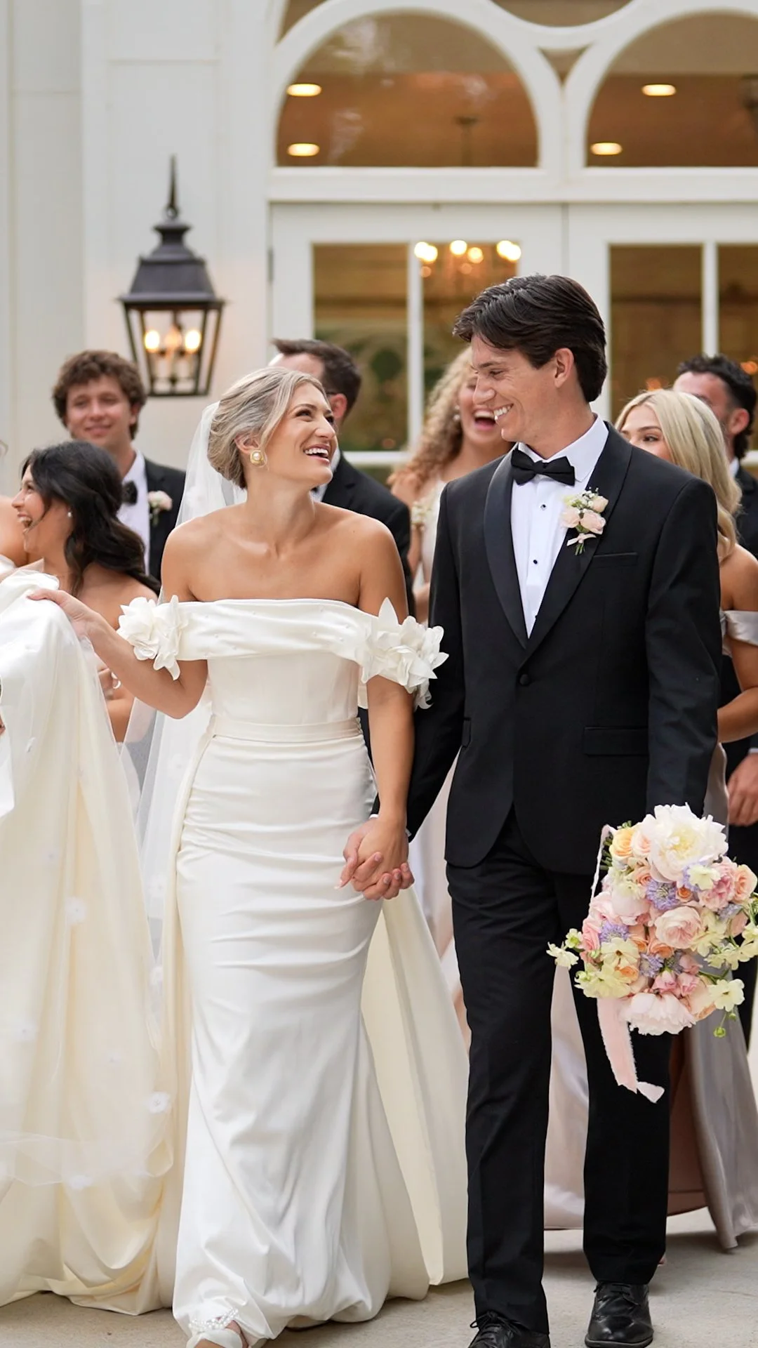 A bride and groom holding hands and smiling at each other during their wedding ceremony, surrounded by wedding guests in formal attire inside a decorated venue.