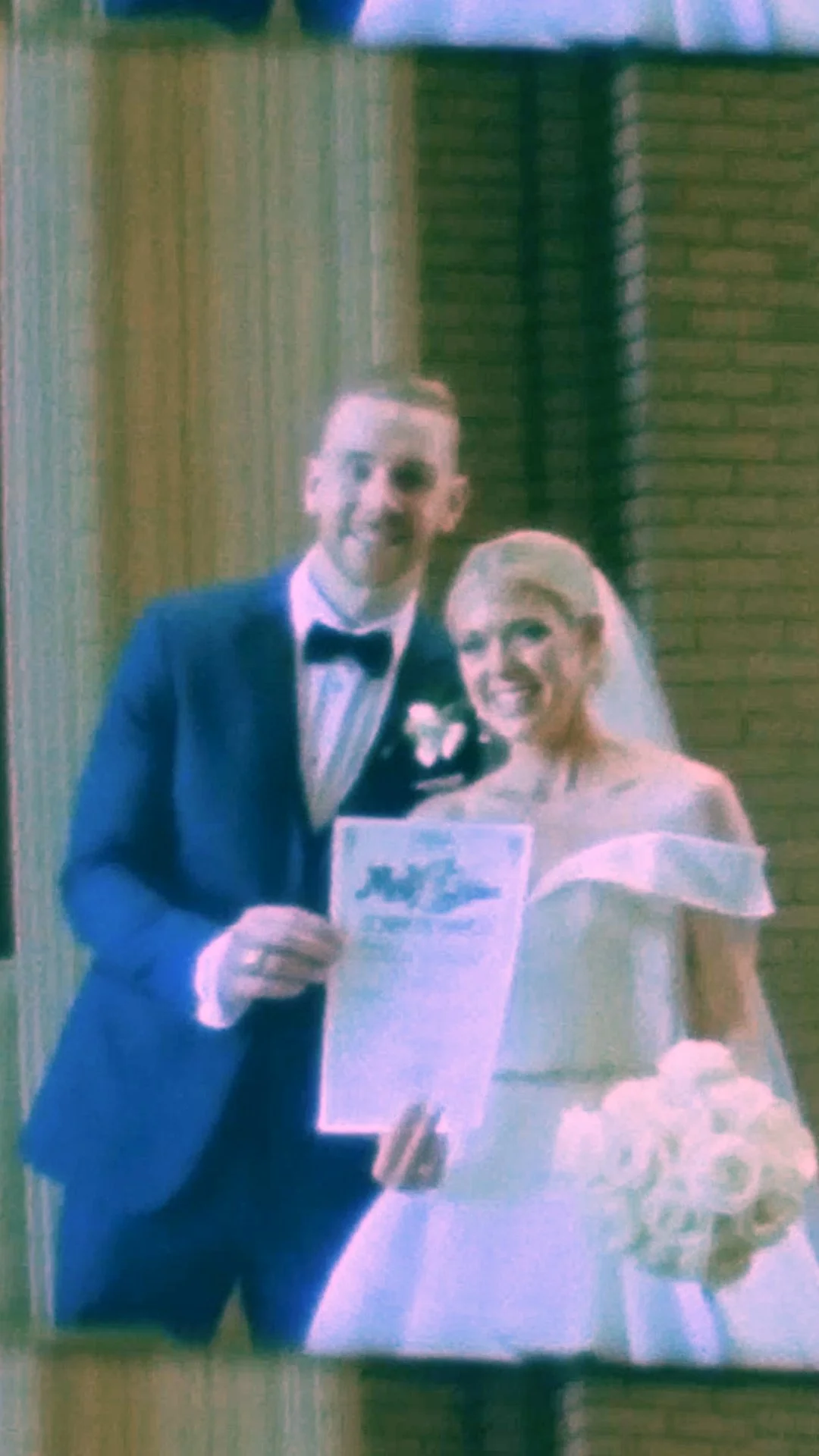 A newlywed couple smiling and holding a wedding bouquet and a wedding photo inside a venue with brick and wooden walls.