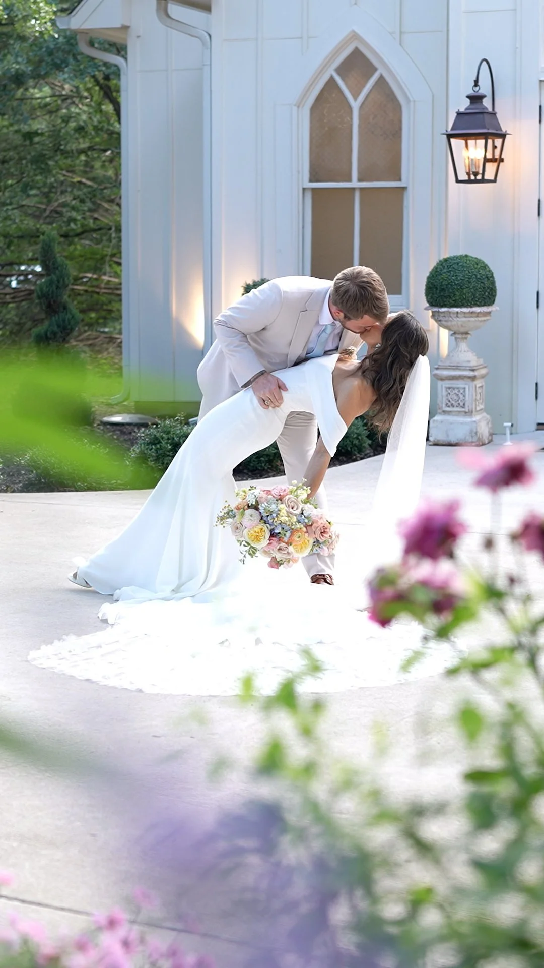 A man and woman dressed in wedding attire, with the man leaning down to kiss the woman who is holding a bouquet of flowers, in front of a church or similar building with a lit lantern outside.