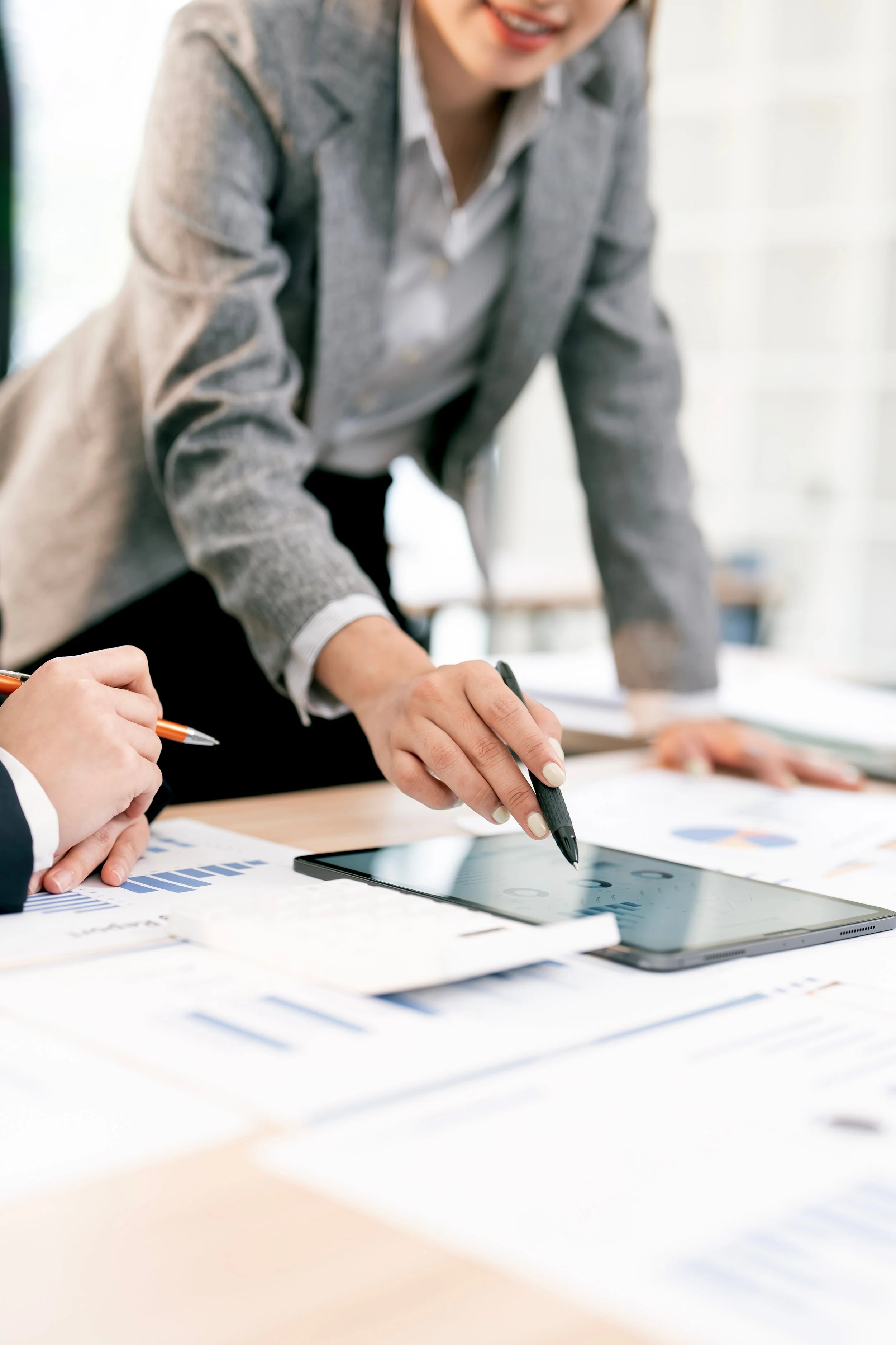 People in business attire working on documents with charts and a tablet on a desk in a bright office.