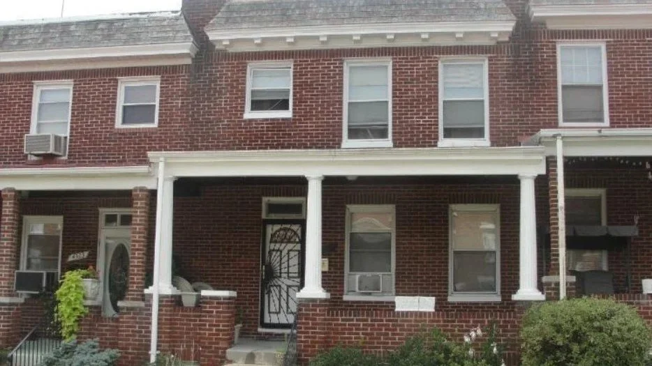The image shows a red brick building with four windows, two of which are visible. The windows have black metal frames and are reflective. There are green leaves from a tree partially covering the upper right corner of the building. Two of the lower windows have signs made of paper with handwritten messages, one says 'HOPE' with some greenery visible through the glass, and the other has messages including 'WE' and 'THIS' written in colorful letters.