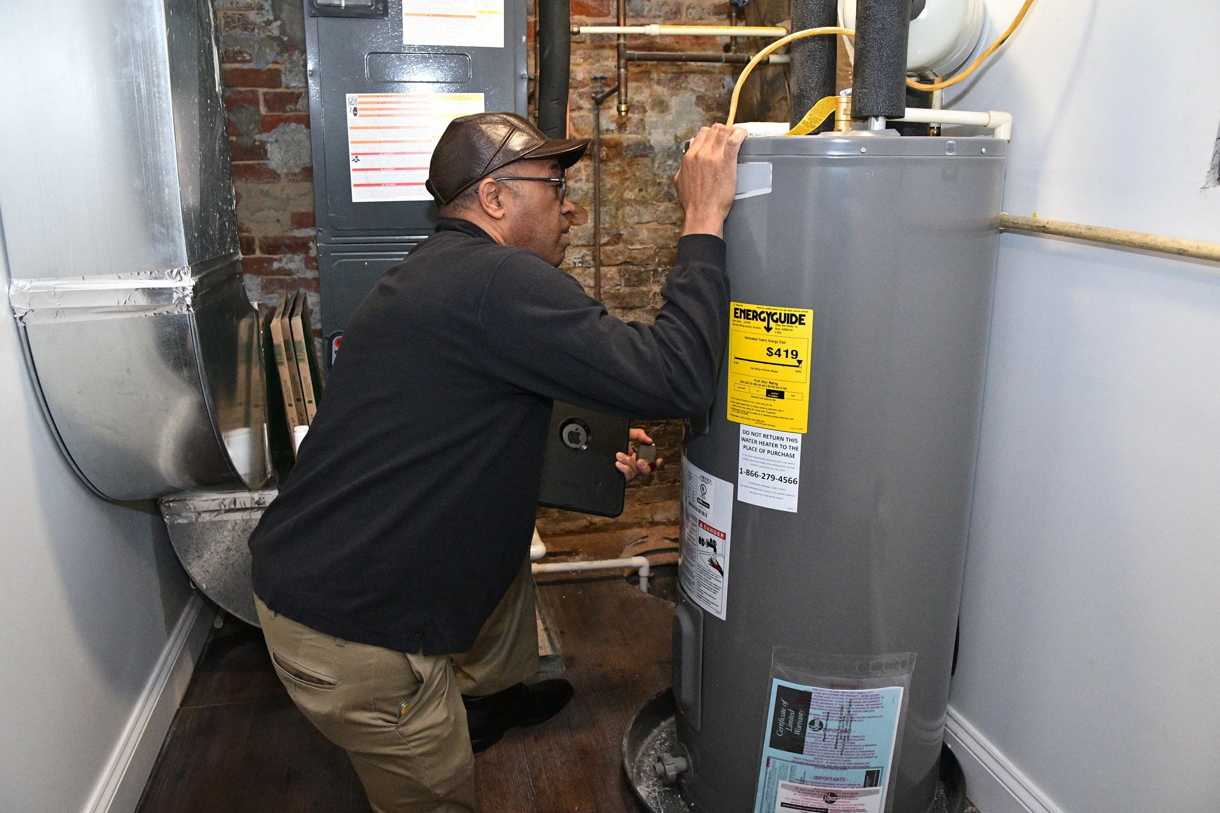 A man wearing glasses, a brown leather cap, a black jacket, and beige pants is kneeling in front of a large gray water heater, inspecting or repairing it in a basement with exposed brick wall and various pipes.