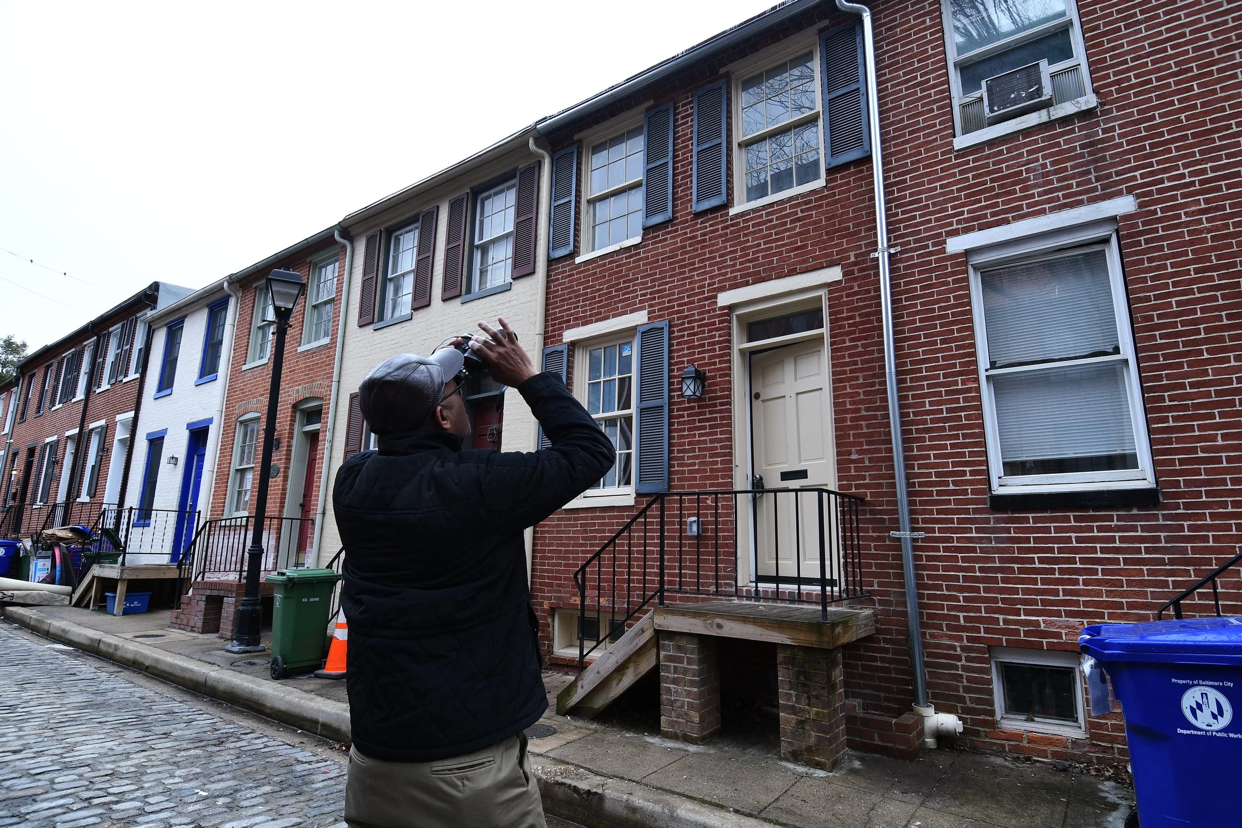 A man wearing a gray cap and black jacket is pointing a camera at a row of brownstone townhouses on a cobblestone street, with one person visible through an open window.