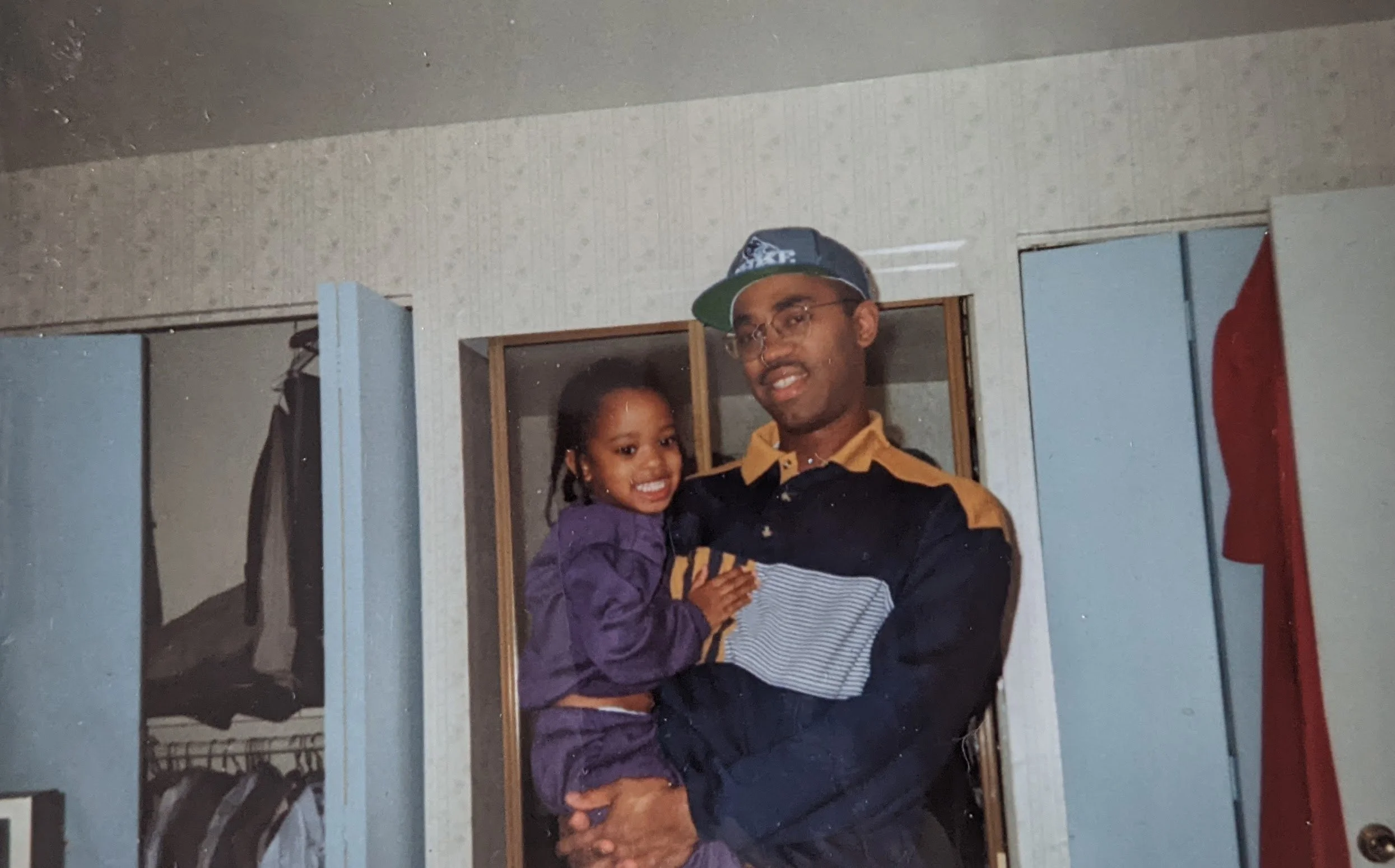 A man wearing glasses, a cap, and a black and yellow sports jacket holds and smiles at a young girl with braided hair, dressed in a purple jacket, inside a room with open closet doors, clothing, and a red and blue item hanging in the background.
