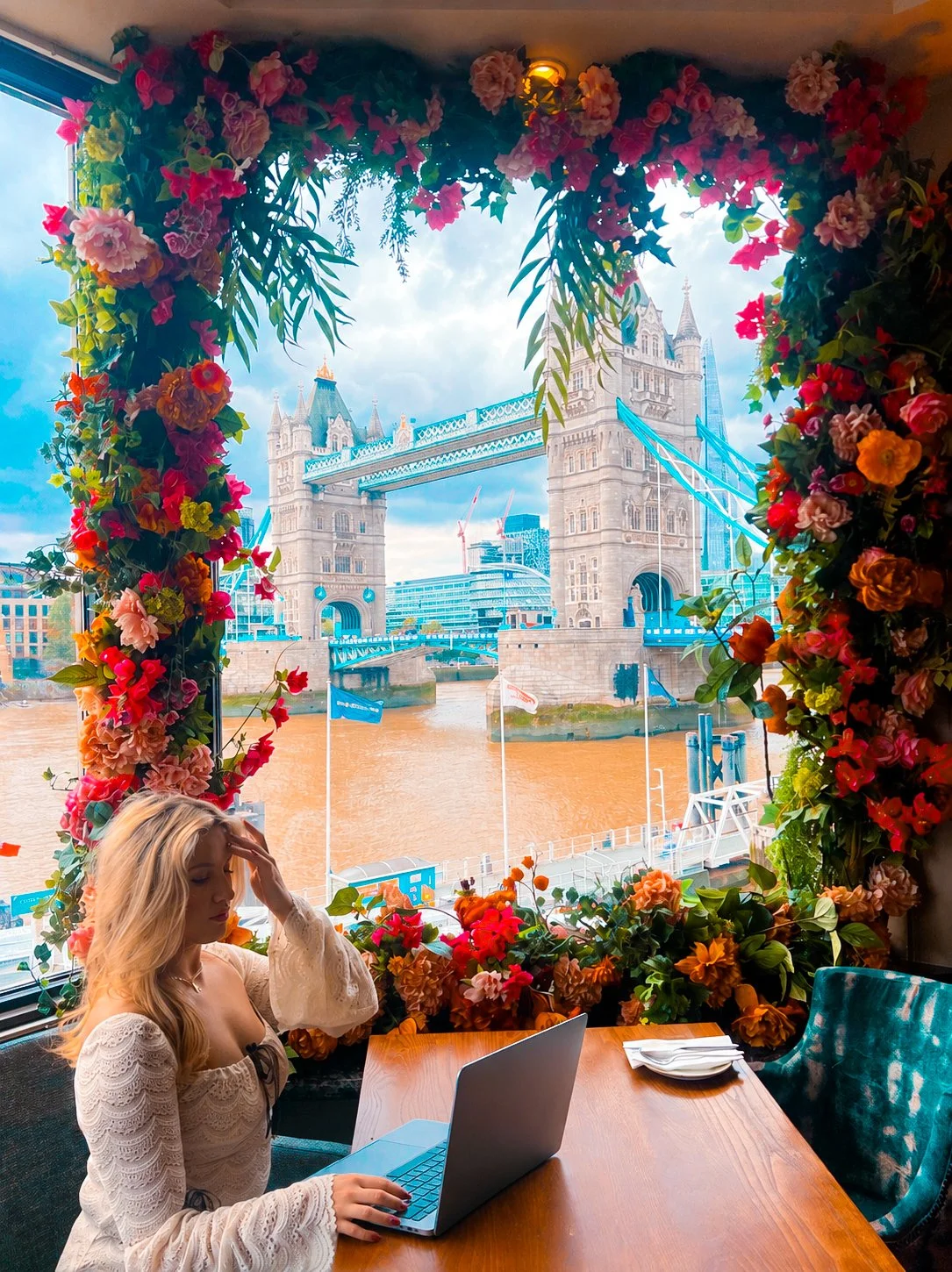 A woman sitting at a wooden table inside a restaurant with a floral window frame, overlooking Tower Bridge in London across the Thames River.