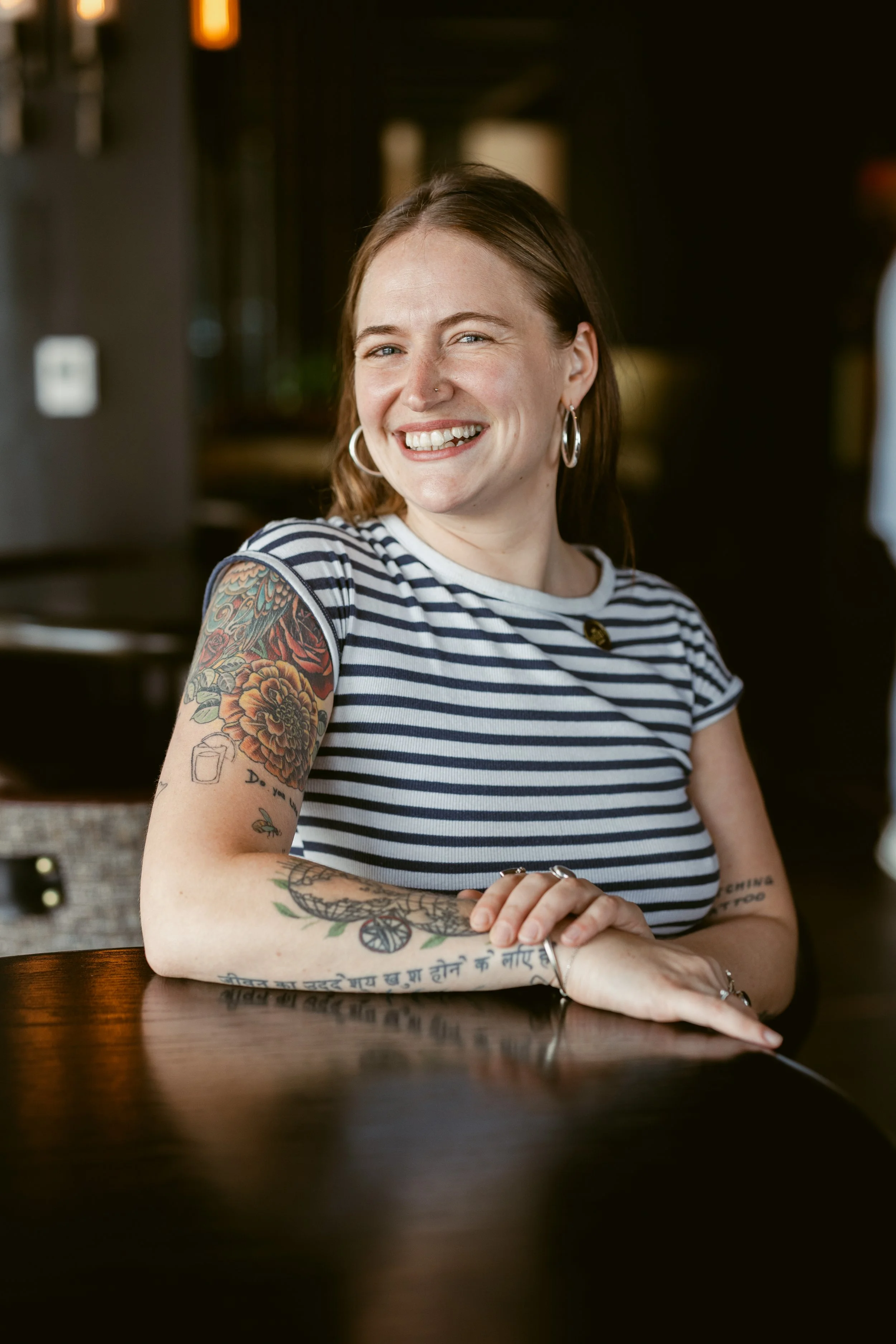 A smiling woman with tattoos and hoop earrings, wearing a striped t-shirt, sitting at a dark wooden table in a restaurant or cafe.