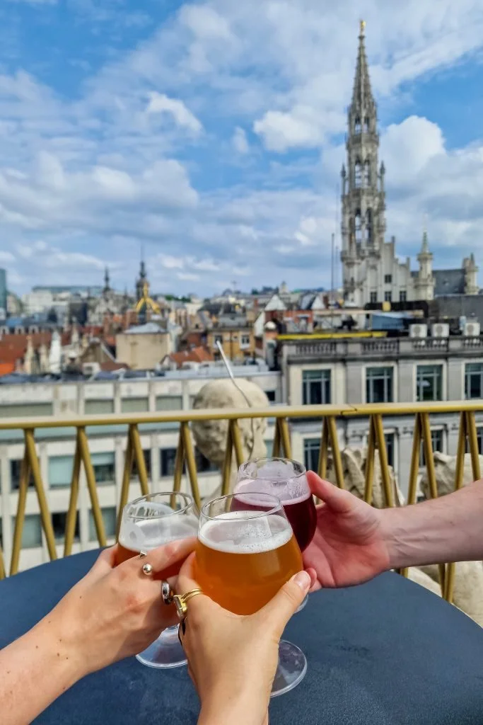 Three people clinking glasses of beer and wine on a rooftop terrace with city buildings and a blue sky in the background.
