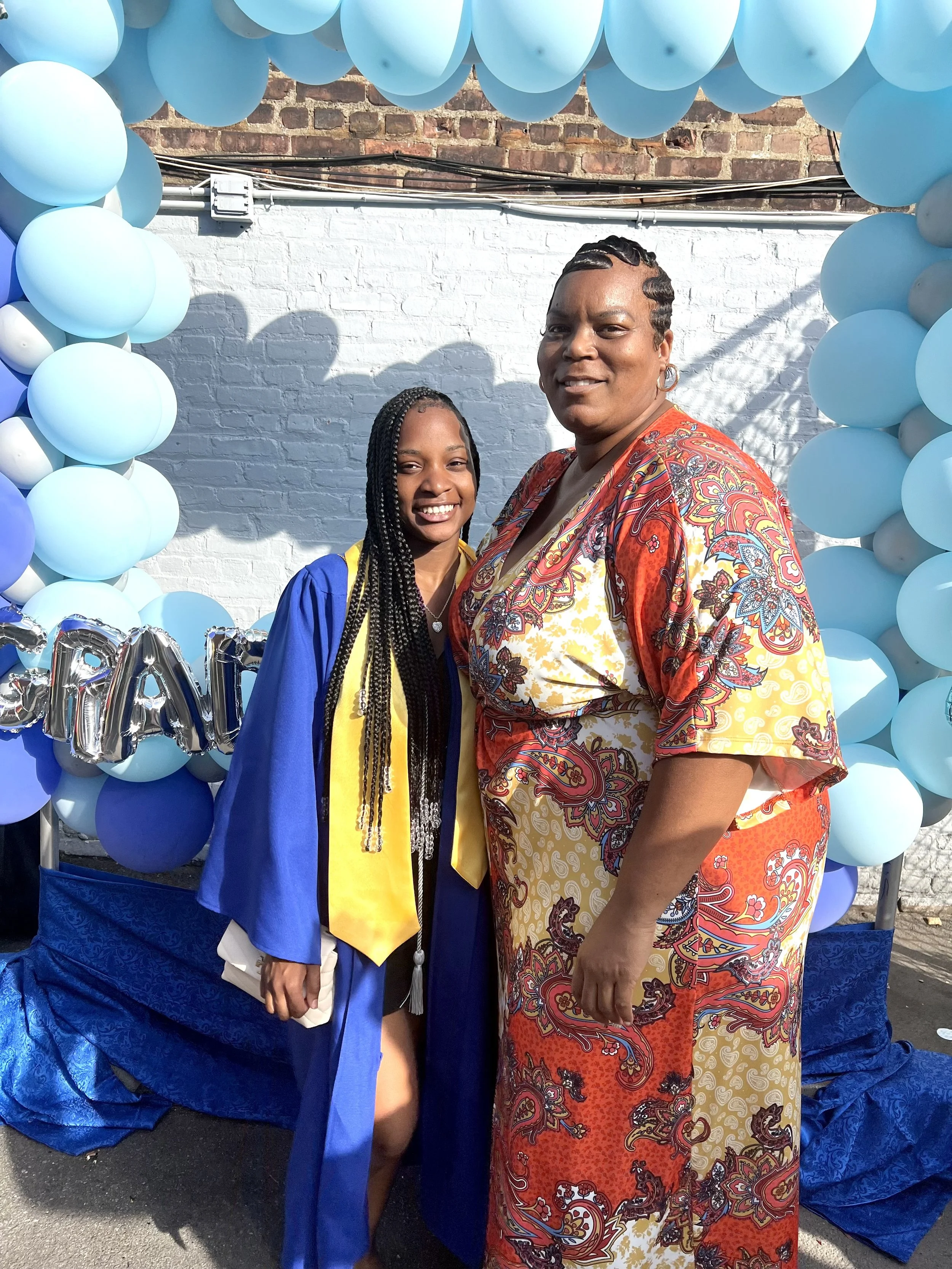 A young woman in a blue graduation gown and yellow stole standing next to a woman in a colorful dress, both smiling, at a graduation celebration with blue and silver balloons in the background.