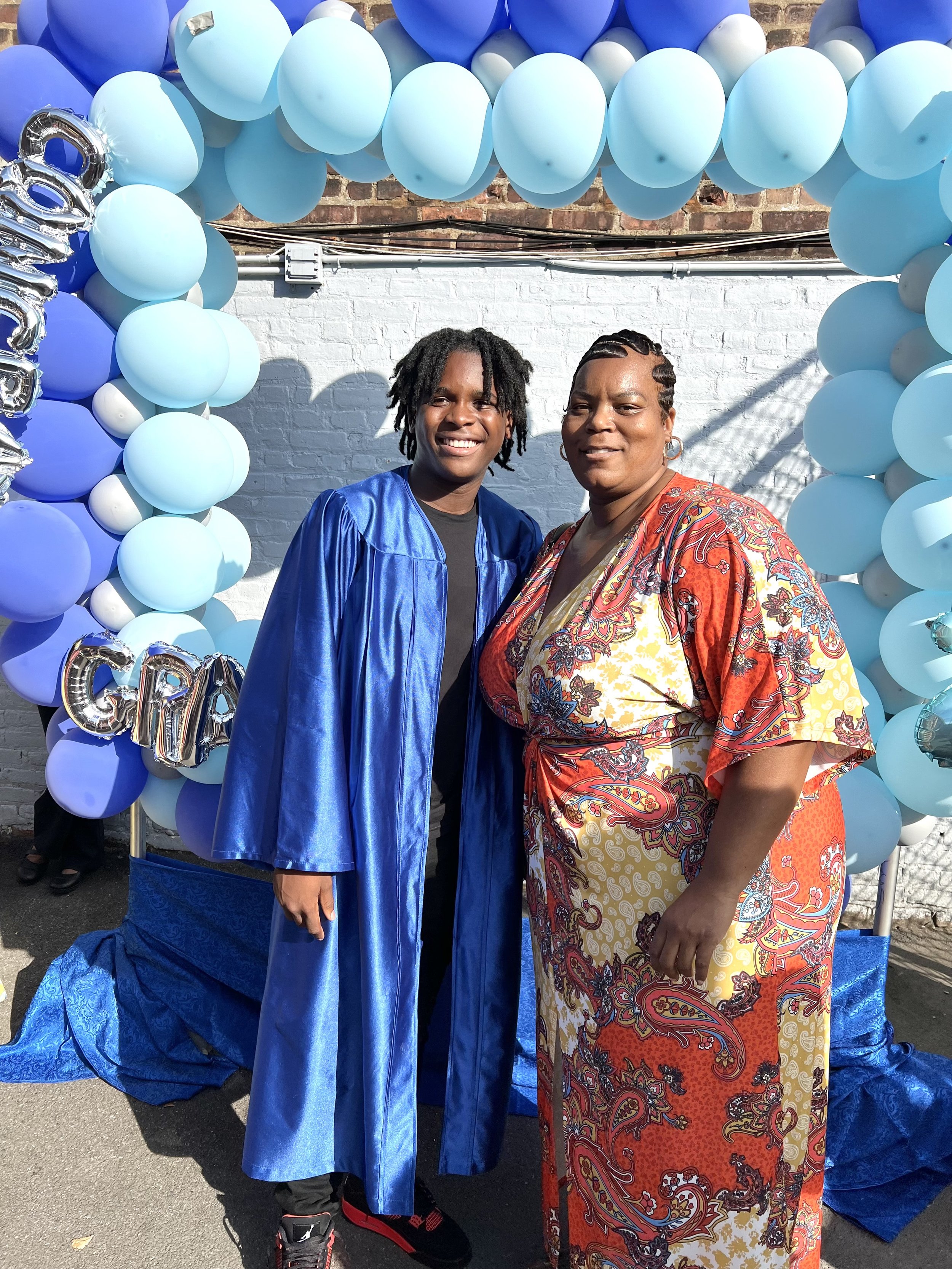 A young man in a blue graduation gown standing next to a woman in colorful clothing, both smiling, under a balloon arch and decorations.