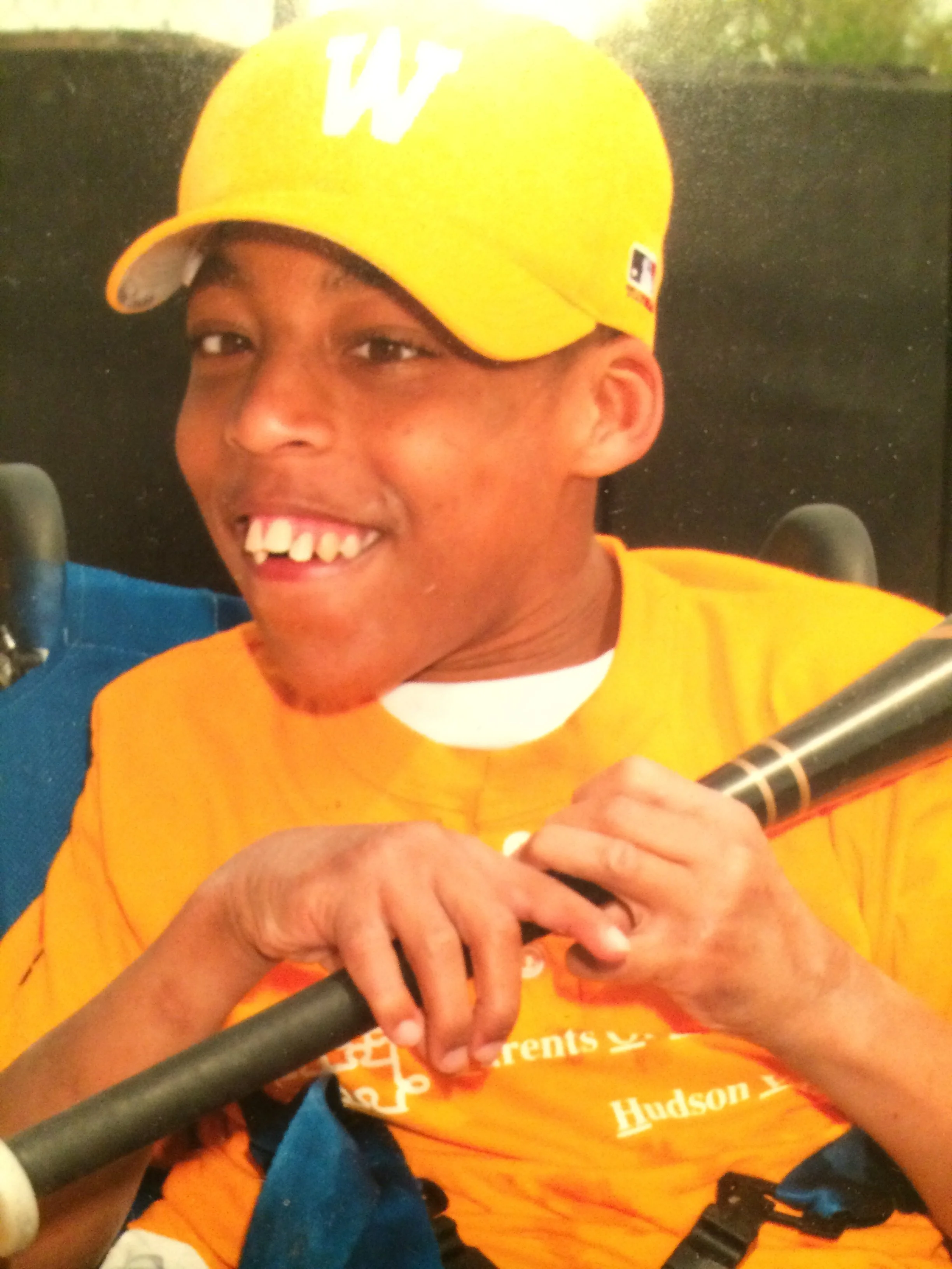 A young boy wearing a yellow baseball cap with a white logo and a yellow and blue sports shirt smiling and holding a microphone.