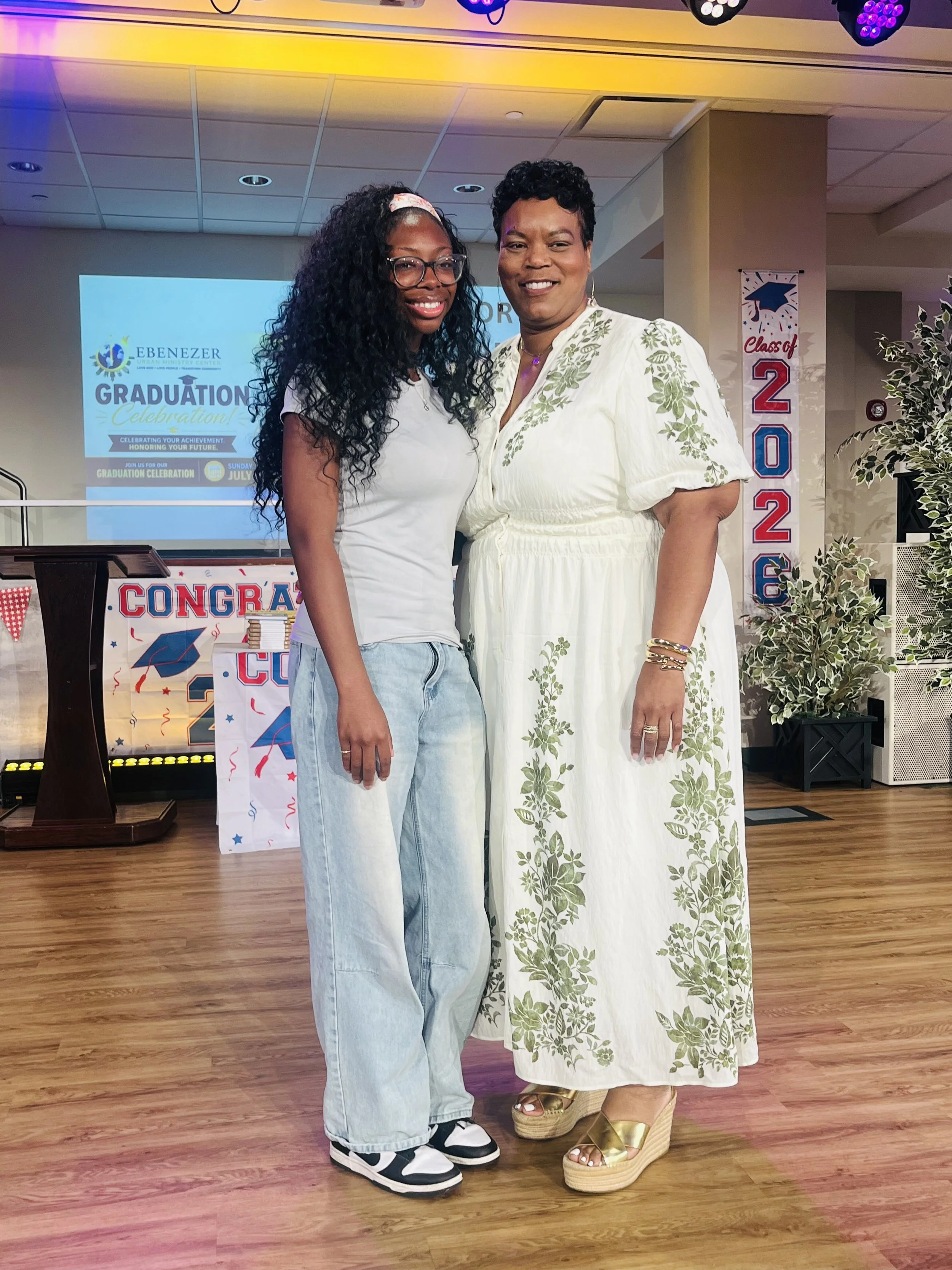 Two women standing together indoors at a graduation celebration, smiling and posing for the photo. One woman has long black curly hair, glasses, and is wearing a white t-shirt and light jeans. The other woman has short curly black hair and is wearing a white dress with green floral embroidery and gold wedge sandals. There is a graduation-themed banner and decorations in the background.