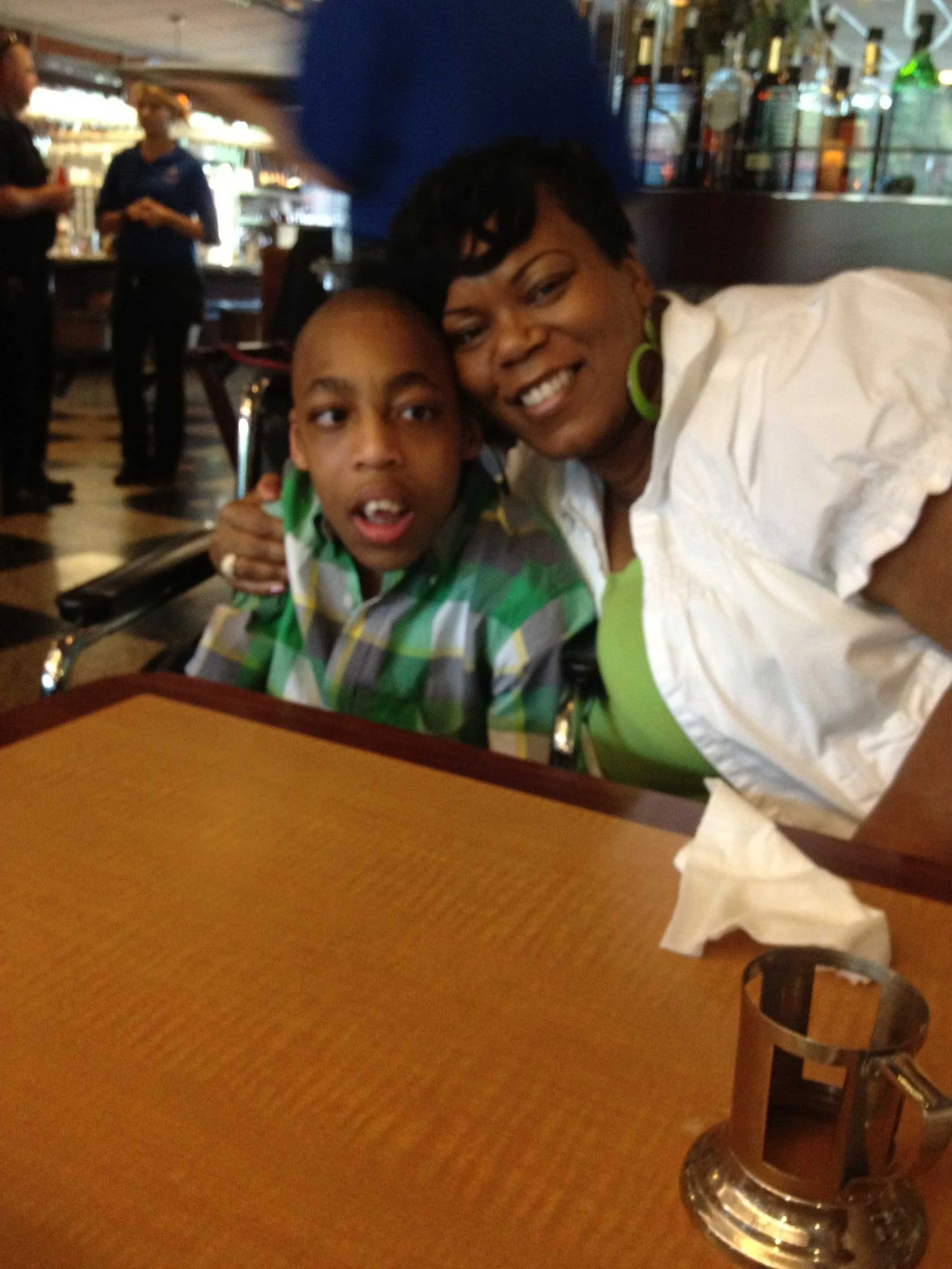 A mother (Rosemary) and her son (Karem) sitting at a restaurant table, smiling and posing for a selfie, with napkins and a napkin holder on the table, in a lively restaurant setting with other patrons and waitstaff in the background.