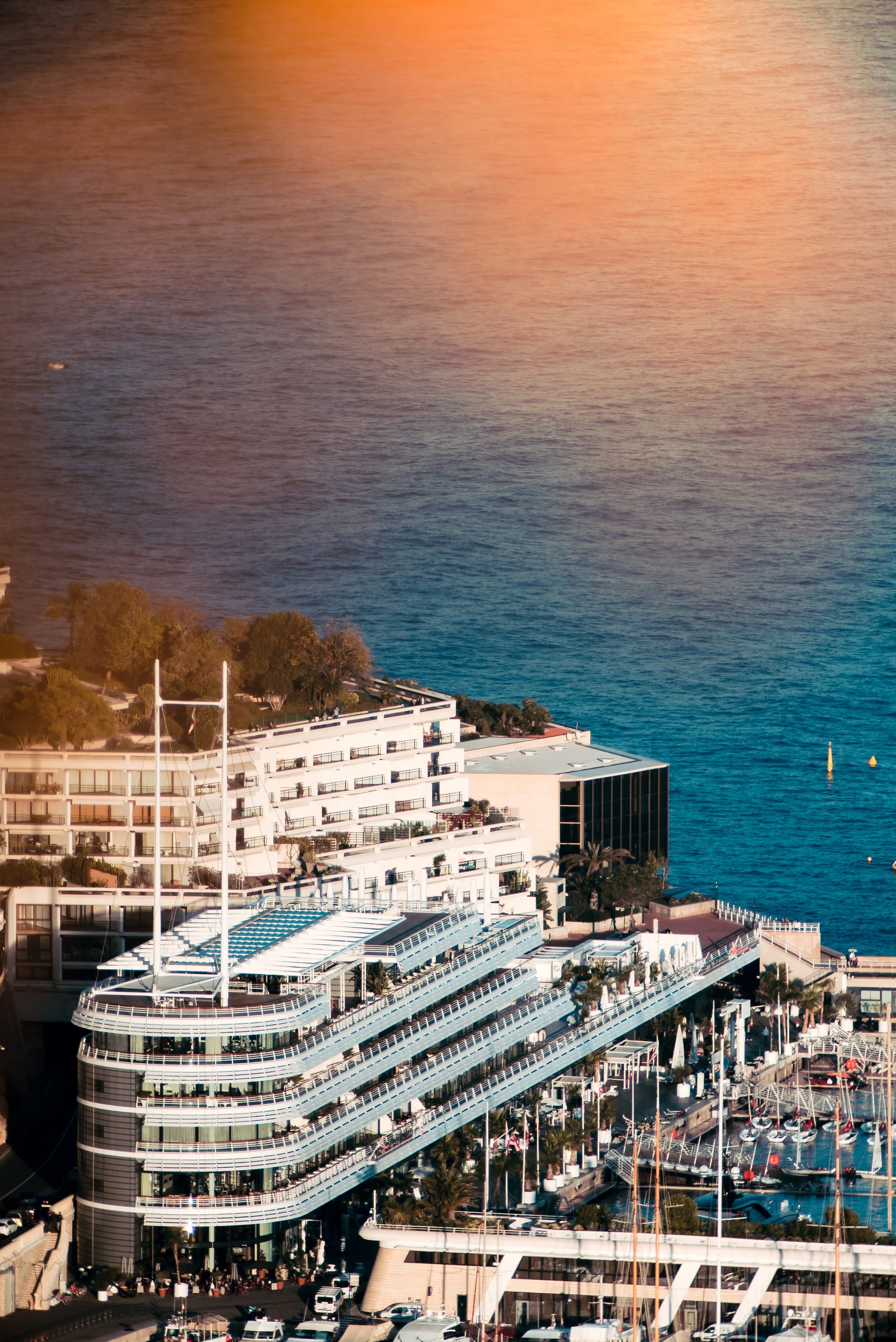 A multi-story building with white exterior and glass balconies near a body of water. The photograph appears to be taken during sunset, reflecting warm orange and pink hues on the water. There are boats and a marina nearby, along with trees and some smaller structures.