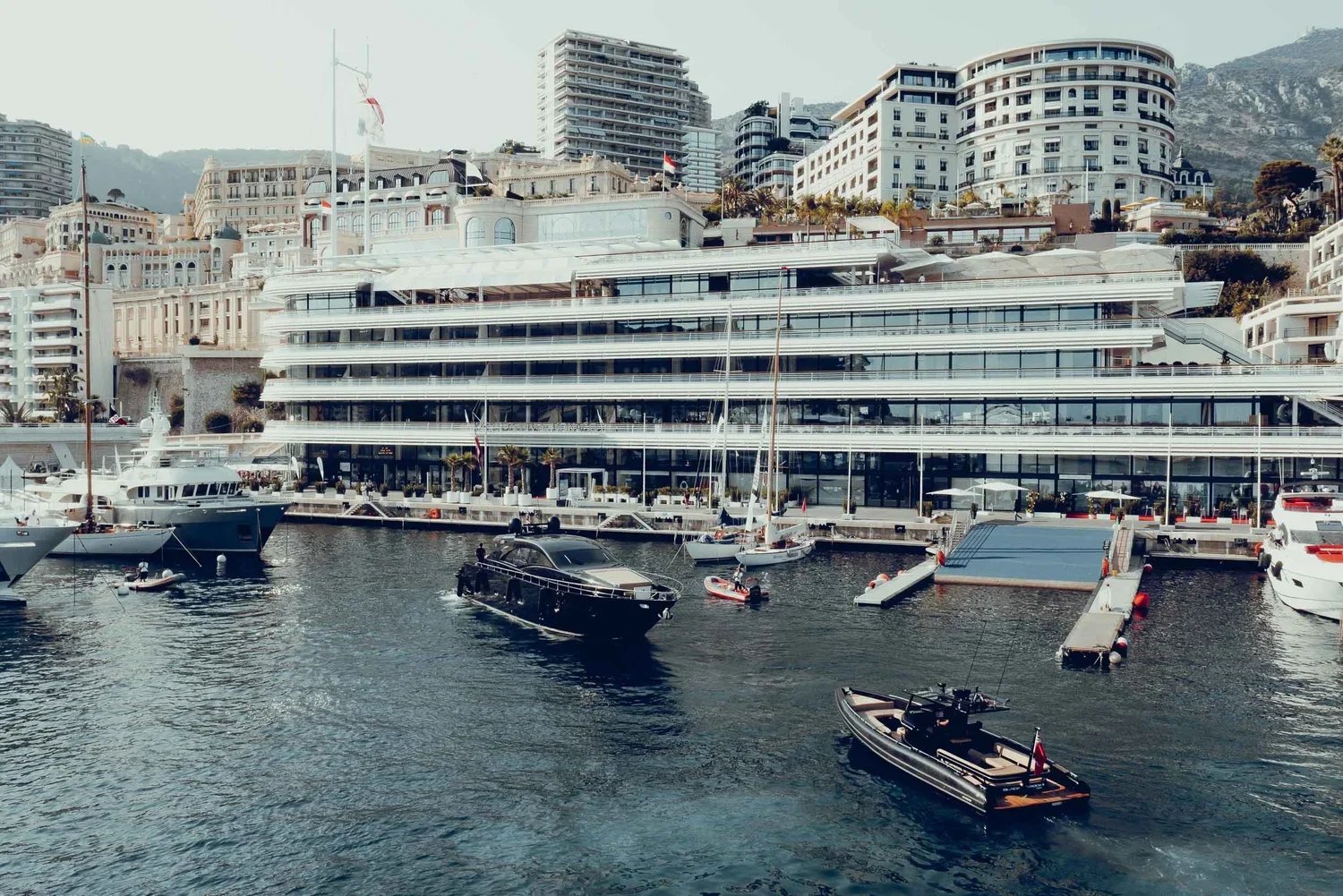 Multiple yachts docked on a marina with a modern building and hillside residential buildings in the background