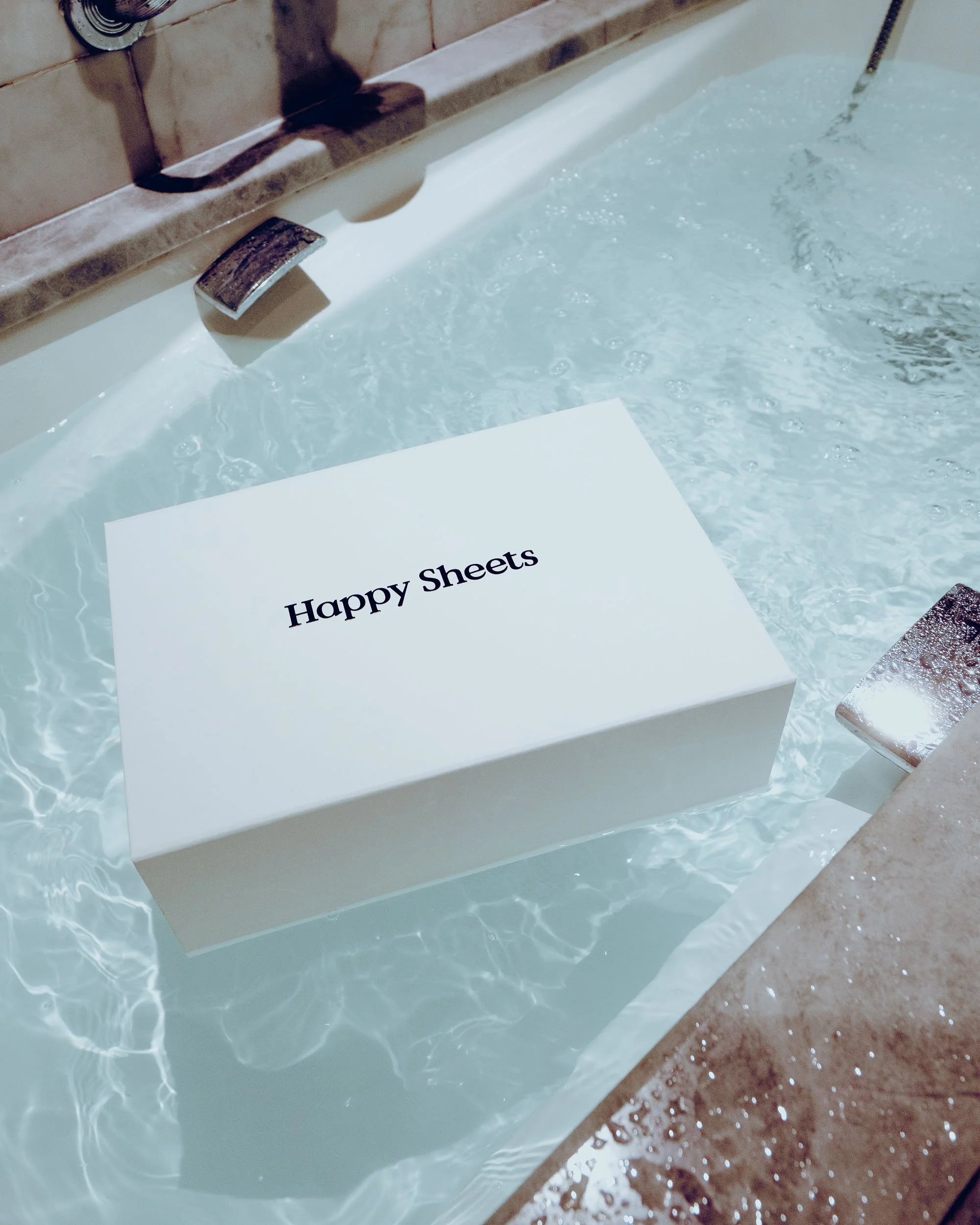 A white box labeled 'Happy Sheets' floating in a swimming pool with clear water, next to a tiled pool edge and a metal pool ladder.
