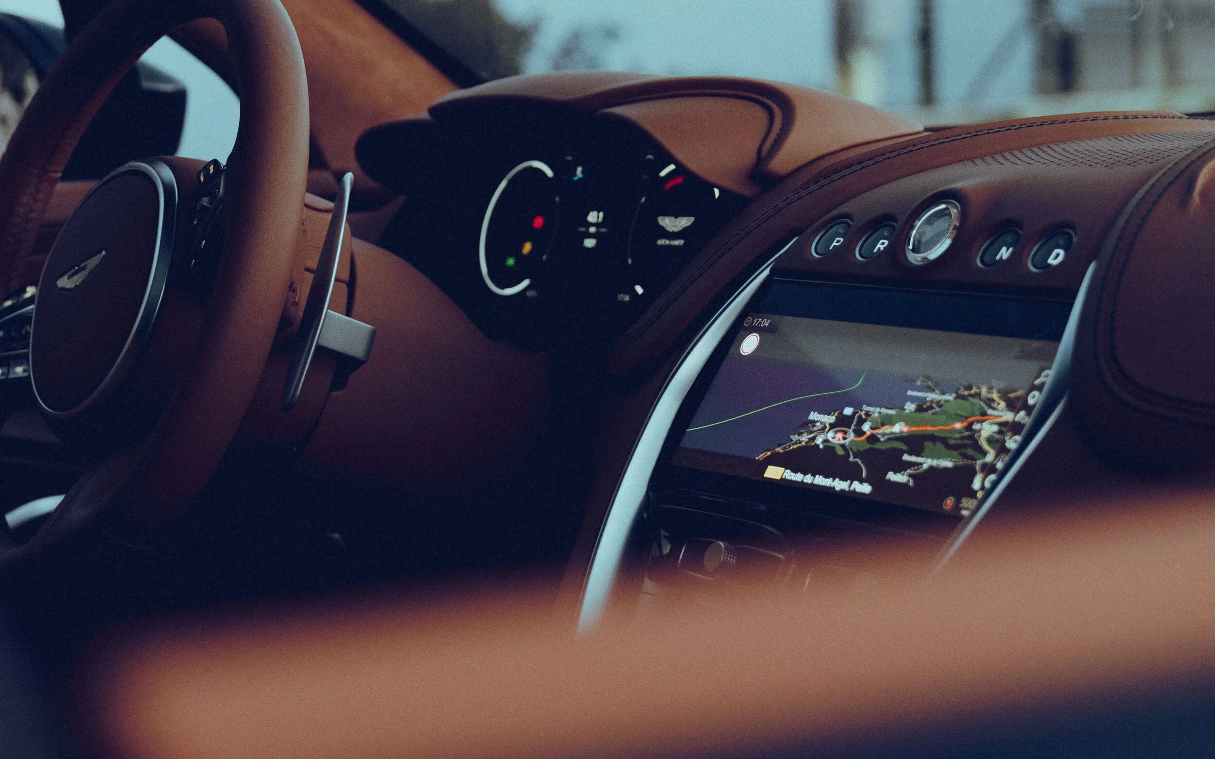 Close-up view of a car's interior dashboard showing an infotainment system displaying a navigation map, gear shift buttons labeled P R N D, and a steering wheel with leather wrapping.