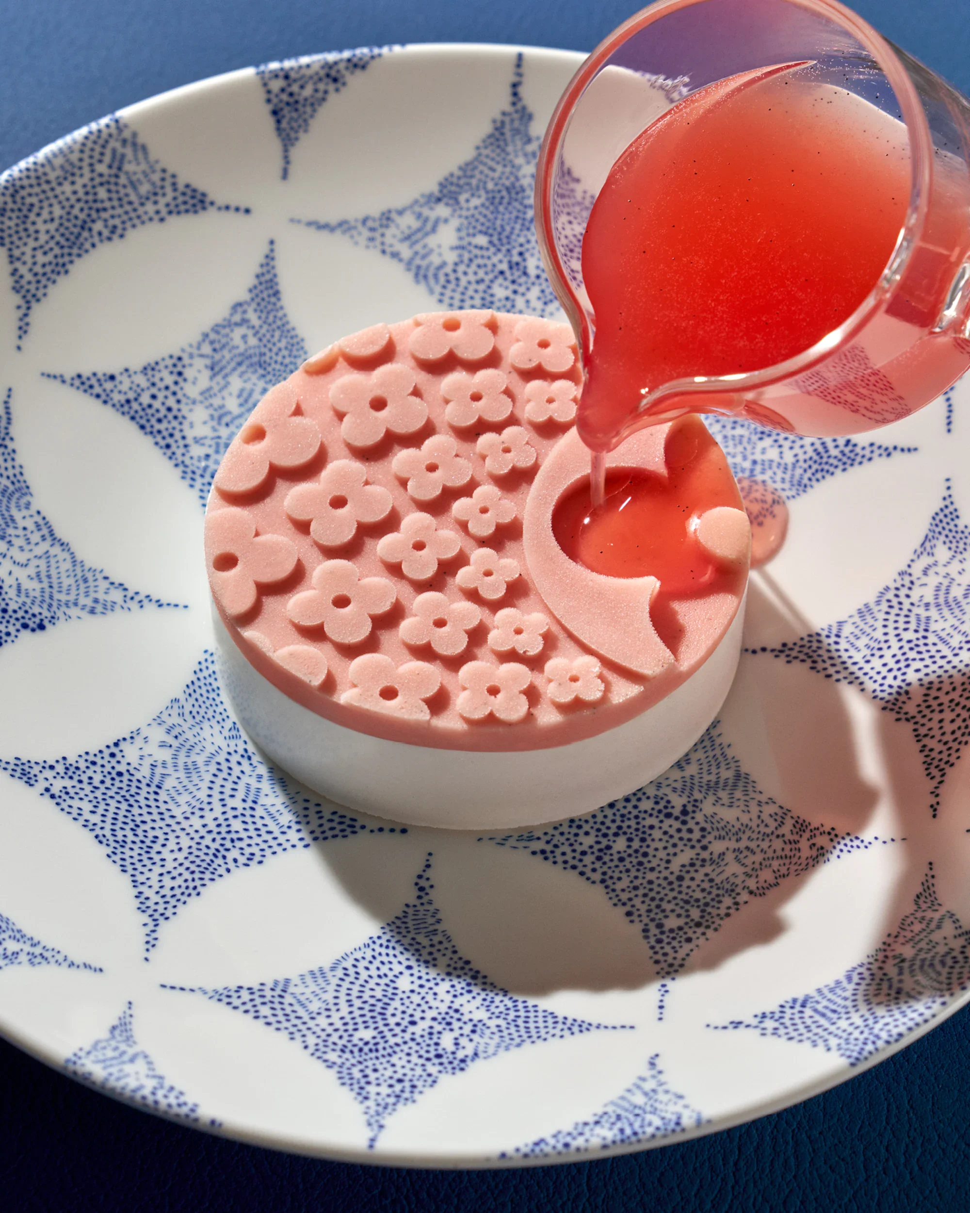 A white bowl with a blue dotted pattern, containing pink soap with a floral design, is placed on a white plate with a blue dotted design, and a pink glass pitcher pours pink liquid soap into the soap.
