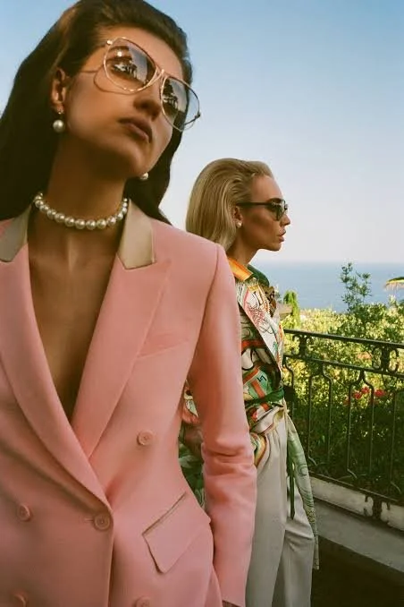 Two women wearing sunglasses and fashionable clothing stand outdoors on a balcony, with greenery and the ocean in the background.