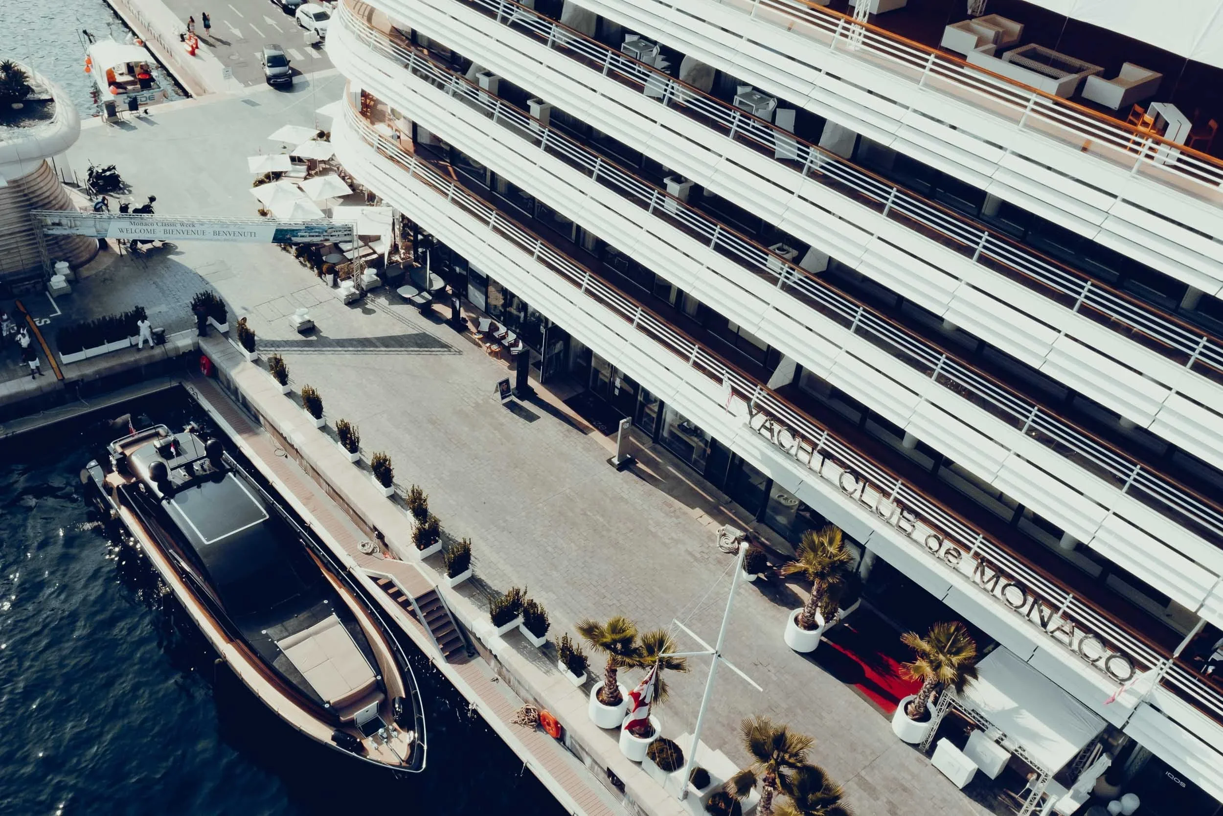 Aerial view of a modern hotel with outdoor patio area, palm trees, and a docked boat in a marina.