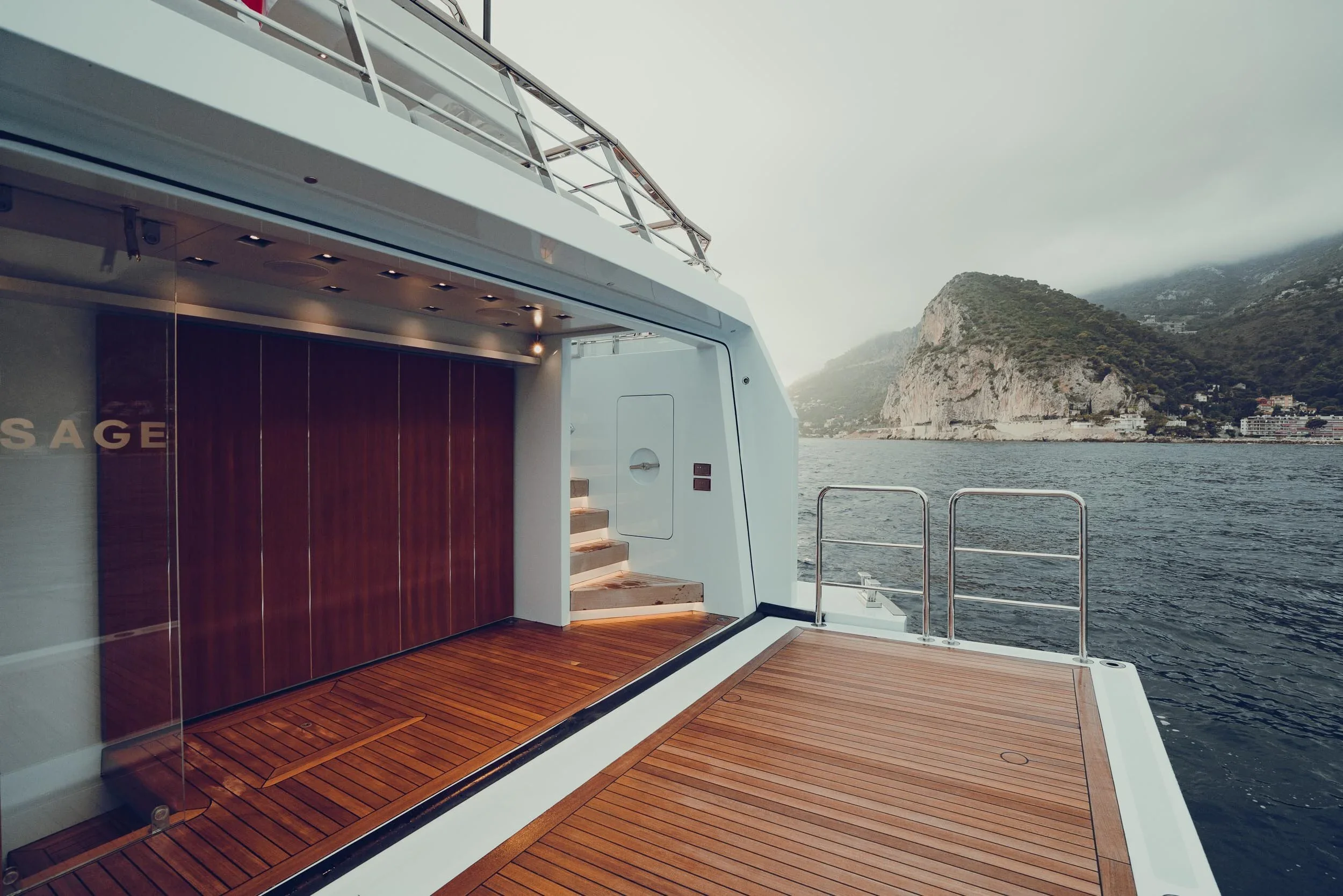 View of the upper deck of a yacht, with wooden flooring and a staircase leading up, overlooking a body of water and mountainous landscape in the distance.