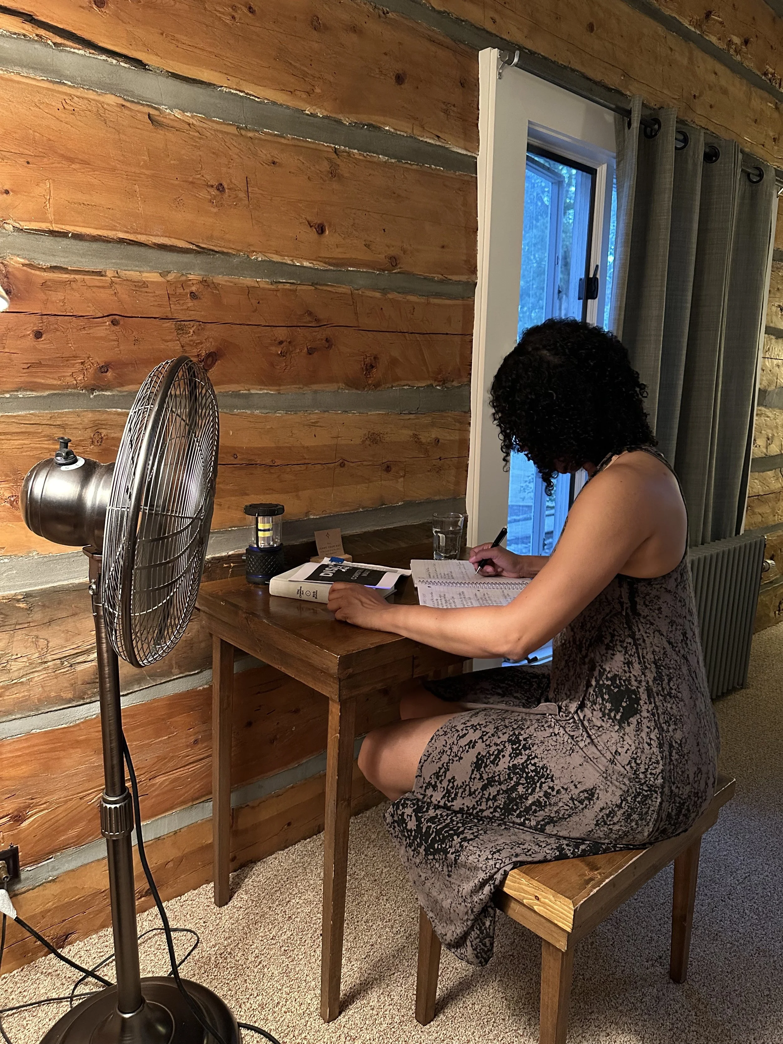 A woman with curly hair wearing a sleeveless dress sitting at a wooden table, writing or reading a notebook, with a fan on a stand next to her, in a room with wooden wall paneling and a window with curtains.