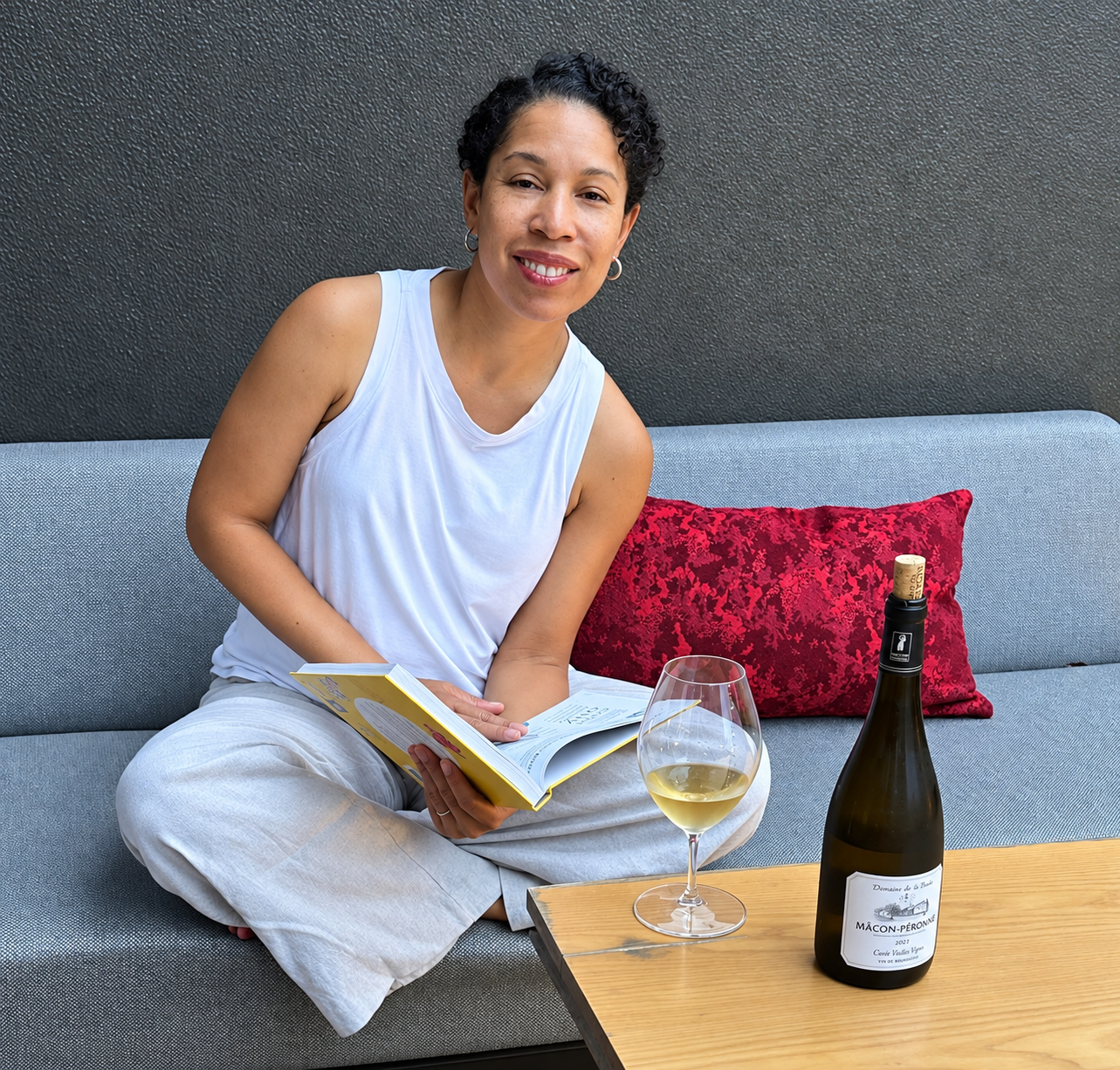A woman with short curly hair sits on a gray sofa, smiling, holding an open book. There is a glass of white wine and a bottle of wine on the wooden table in front of her, with a red decorative pillow behind her.