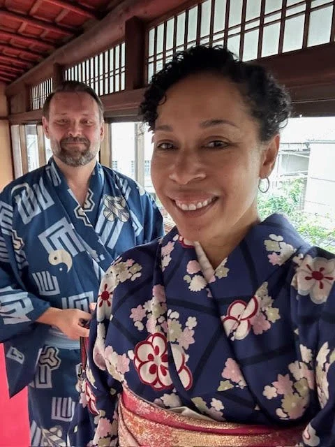 Two people wearing traditional Japanese yukatas, smiling and taking a selfie indoors with wooden windows in the background.
