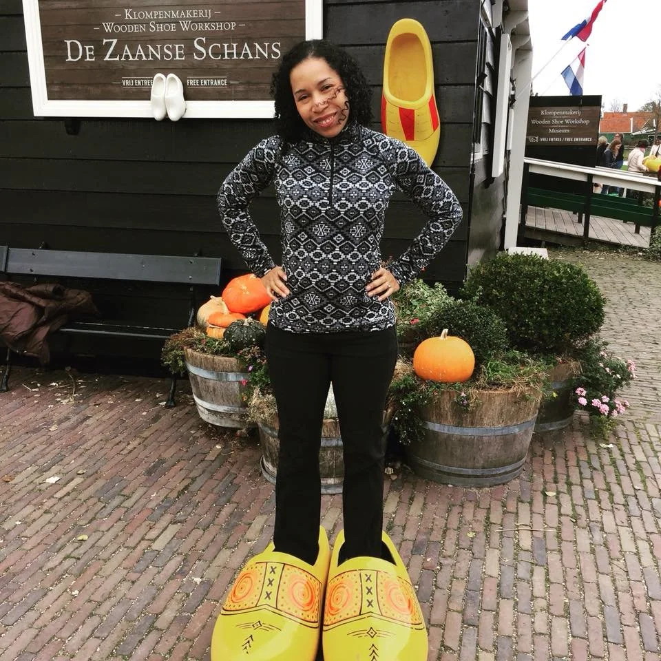 A woman standing outside in front of a black wooden building, smiling, with her hands on her hips. She is wearing a black and white patterned sweater and black pants. She has curly black hair. There are pumpkins and plants in large wooden planters behind her. She is trying on a pair of oversized yellow clown shoes as a joke.