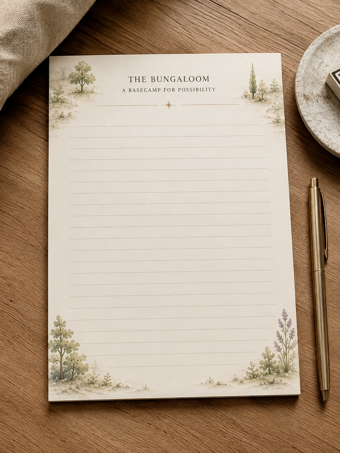 A cream-colored flower and green leafy branch next to a cream-colored notepad with green text and a small plant illustration. The notepad reads 'THE BUNGALOOM, A B&B FOR THE INNER LIFE'.