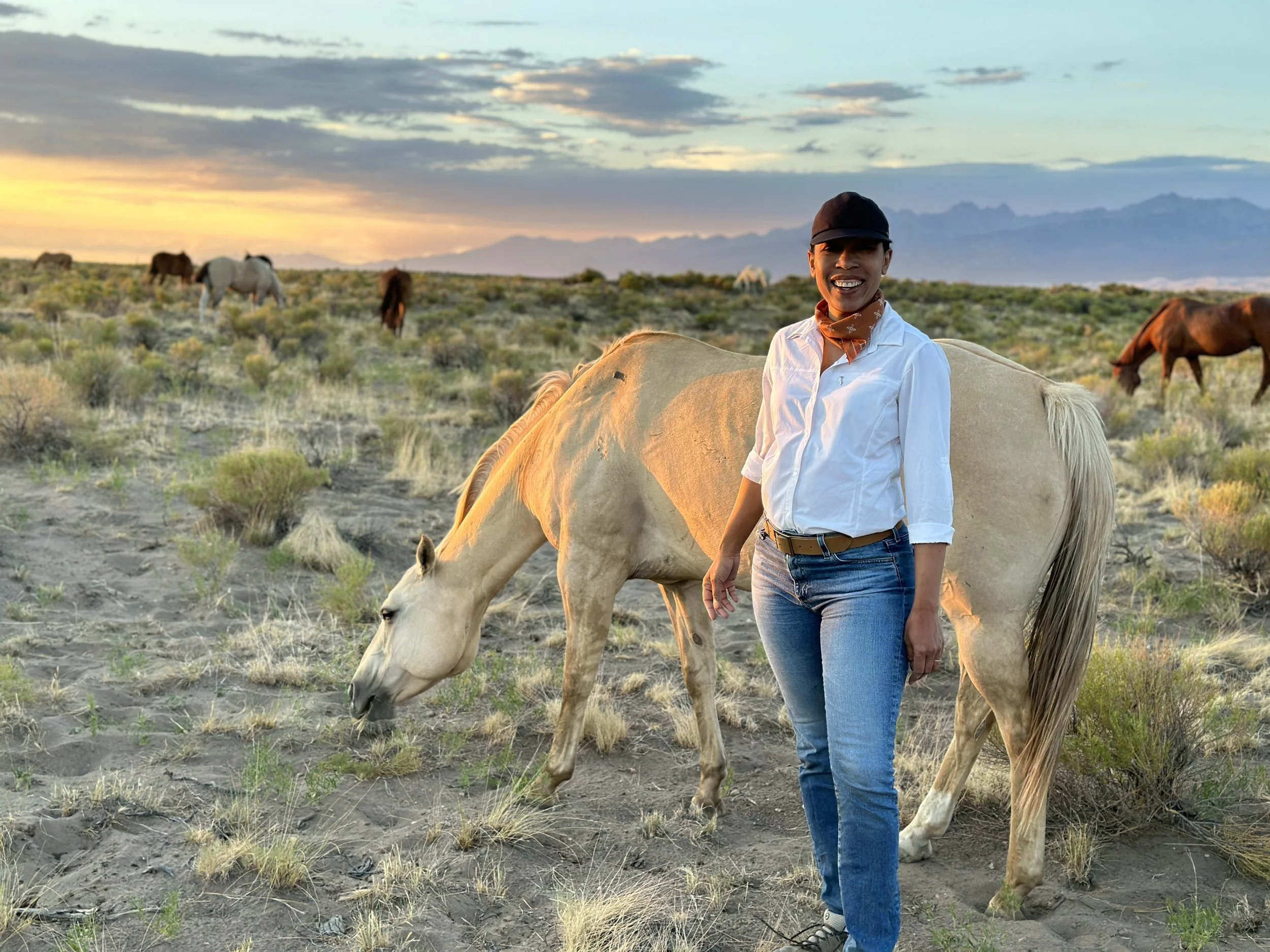 A woman standing next to a white horse in a desert landscape during sunset, with other horses grazing in the background and mountains in the distance.