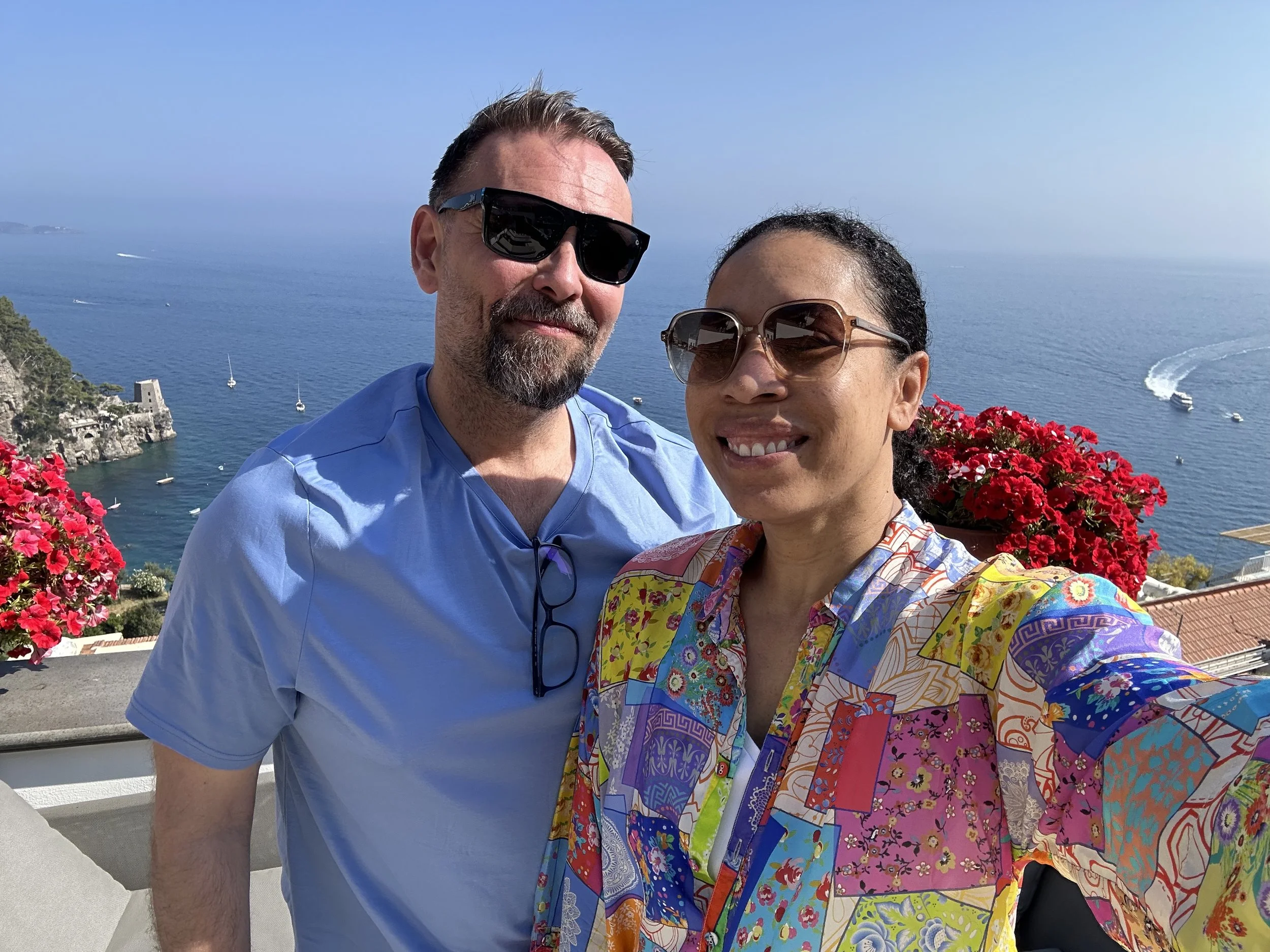 A man and woman taking a selfie together with a scenic ocean view and boats in the background. The man is wearing sunglasses and a light blue shirt, and the woman is wearing sunglasses and a colorful, patterned shirt. They are standing outdoors on a sunny day near a waterfront with pink flowers in the foreground.
