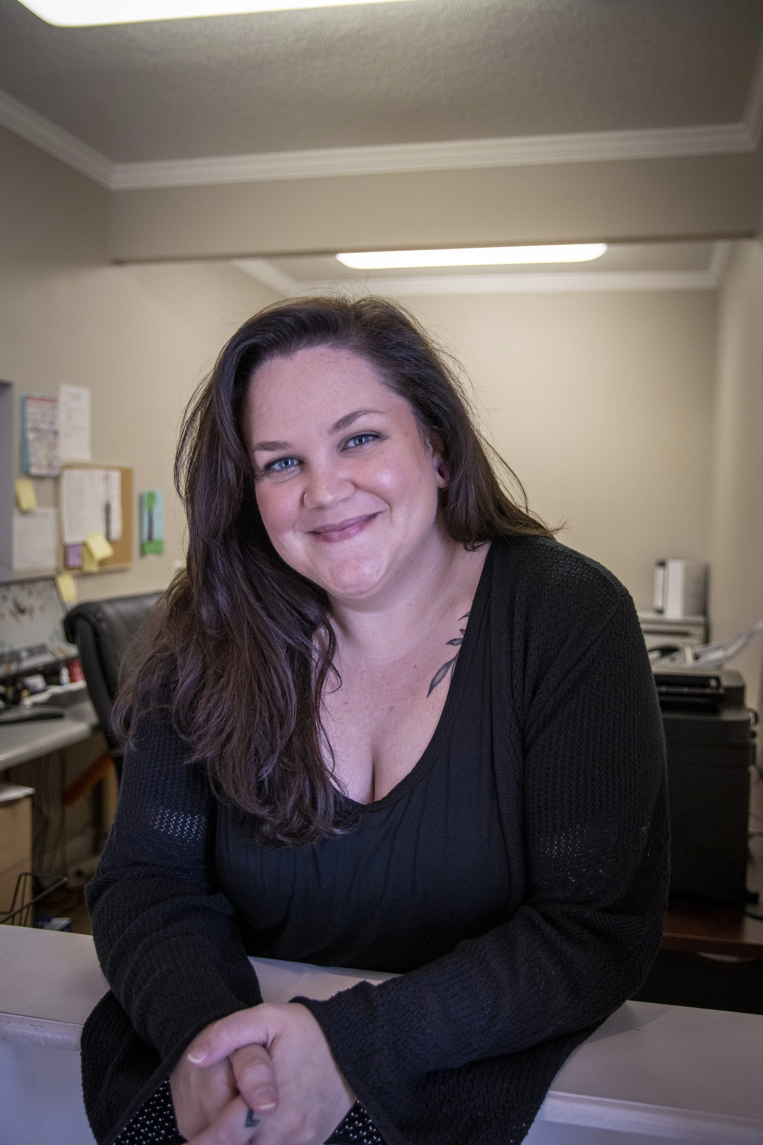 A woman with long, dark hair, blue eyes, and a nose piercing smiling at the camera, sitting at a table in an office setting.