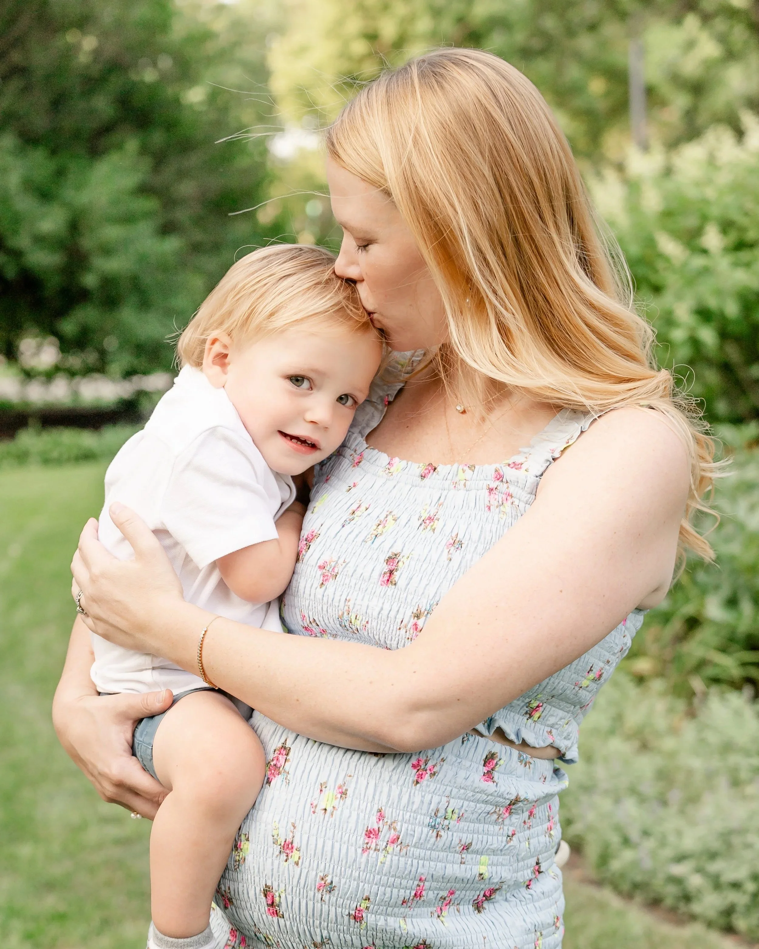 A woman with long blonde hair embracing a young boy with blonde hair in a park with green trees in the background. The woman is kissing the boy on the forehead located in Lincoln Park Chicago.