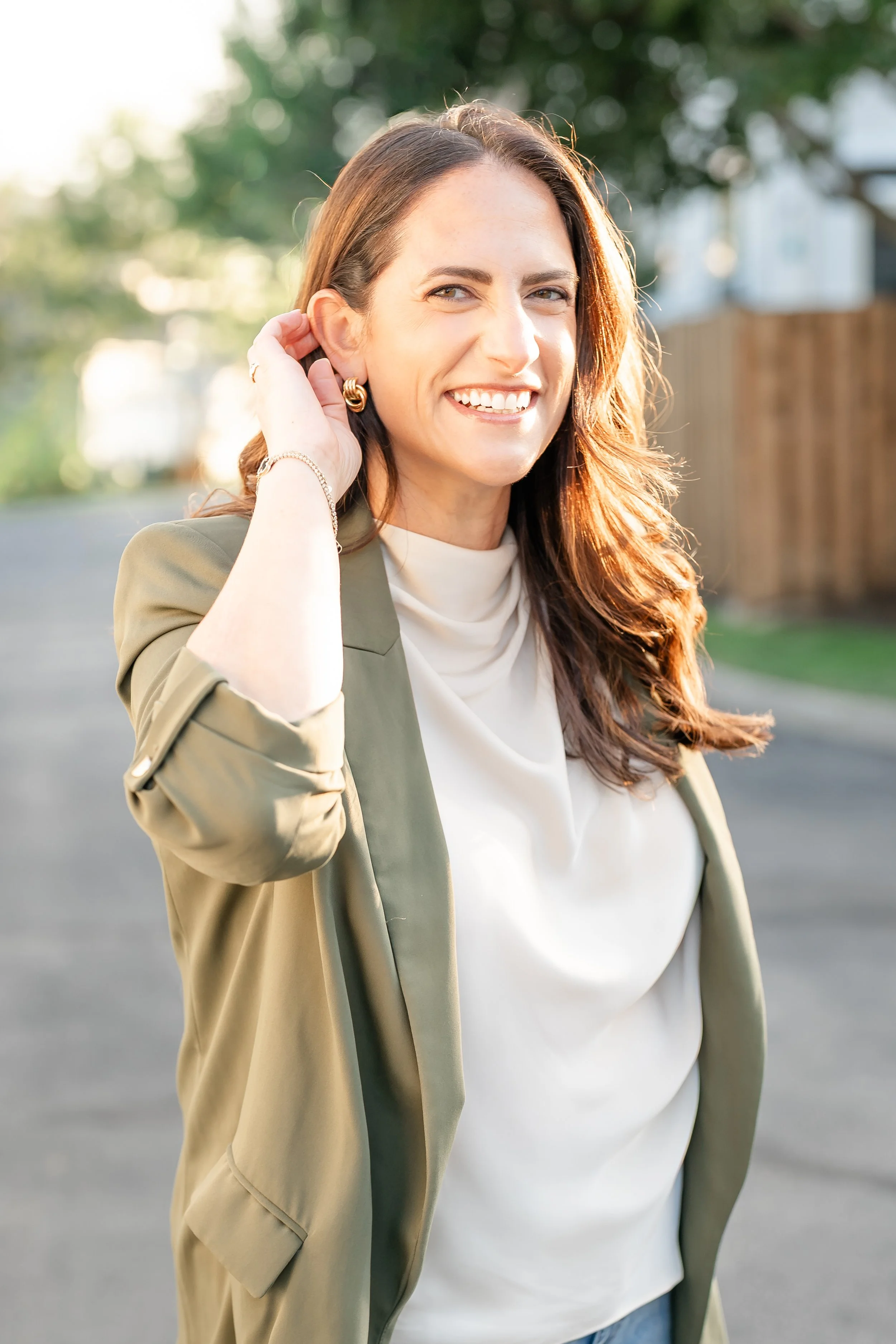 A woman with long brown hair smiling outdoors, wearing a beige blazer and white top, with trees and a house in the background in Glenview IL