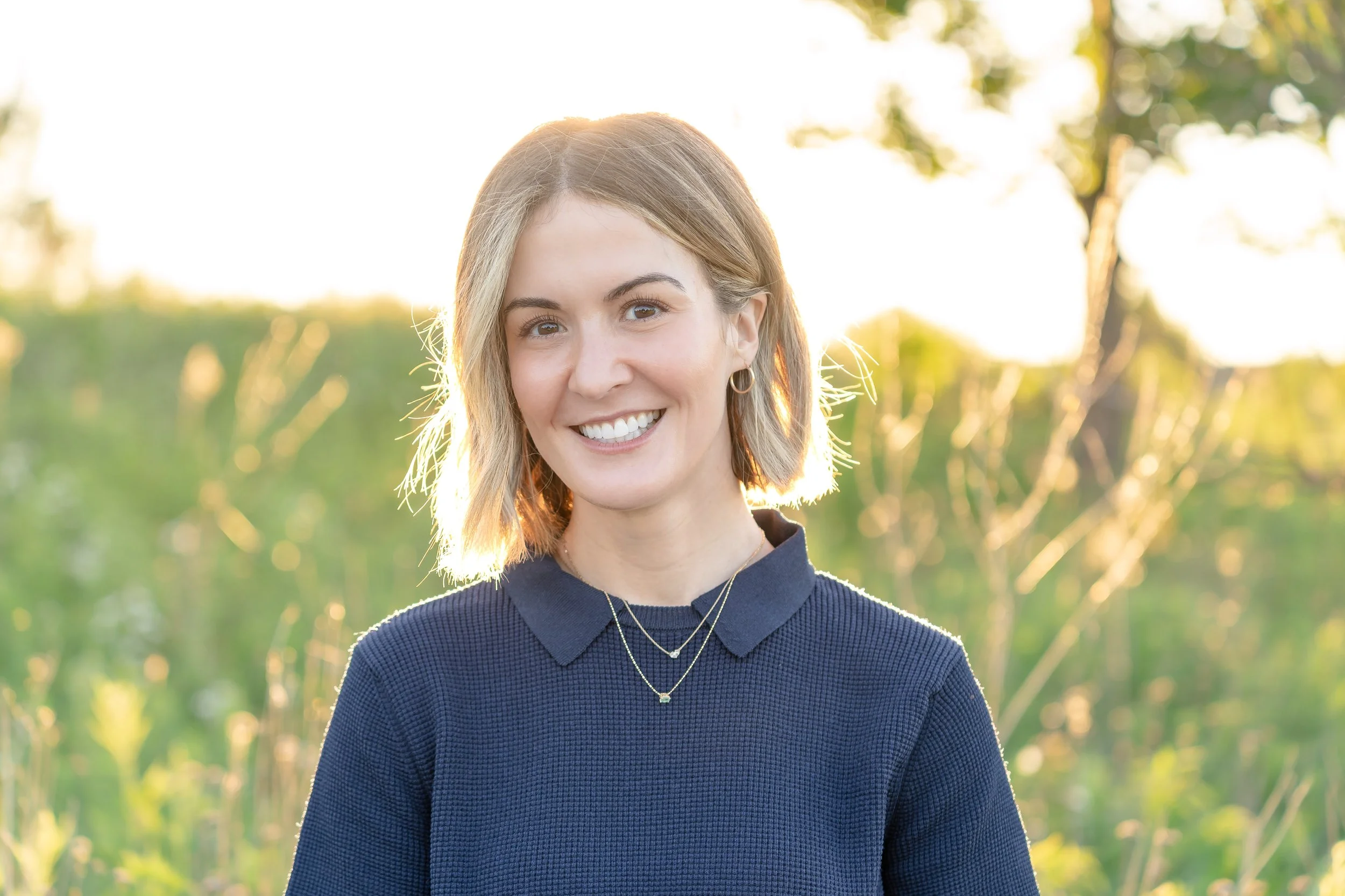 A young woman with shoulder-length light brown hair smiling outdoors during golden hour, wearing a dark blue textured sweater and layered necklaces, with trees and sunlight in the background in Glenview IL.